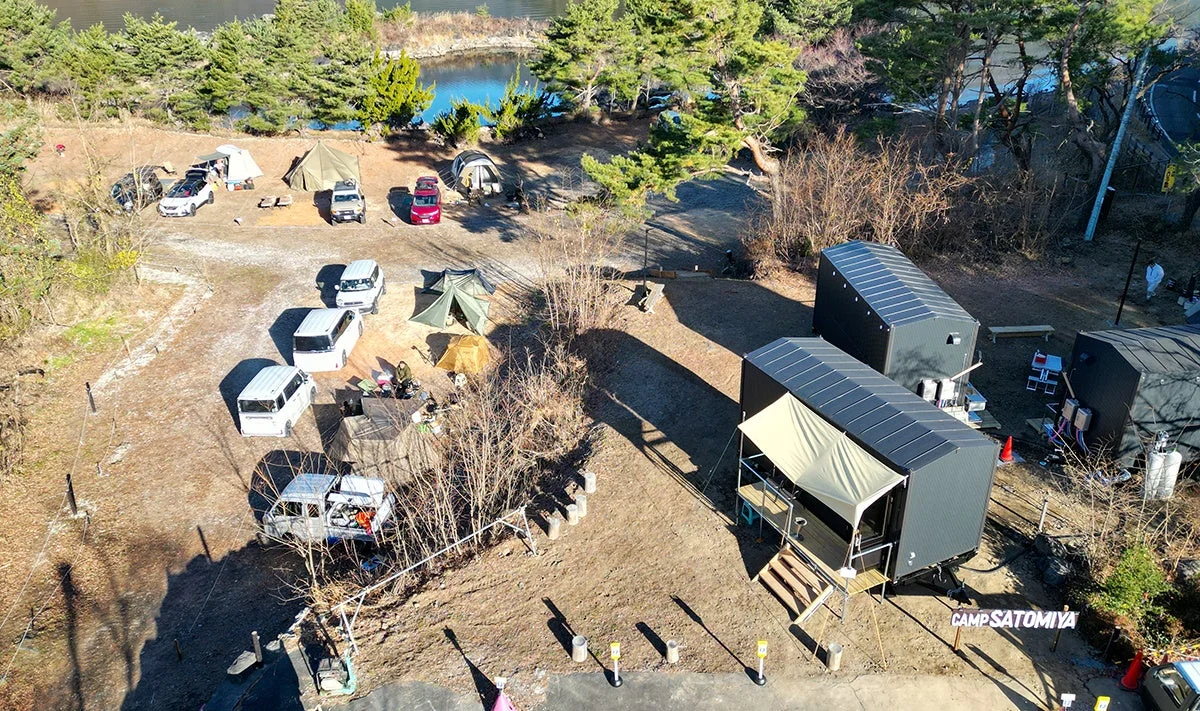 湖畔に広がるキャンプ場の空撮写真。複数のテントや車、モダンなキャビンが点在し、自然の中でアウトドアを楽しむ様子がうかがえます。