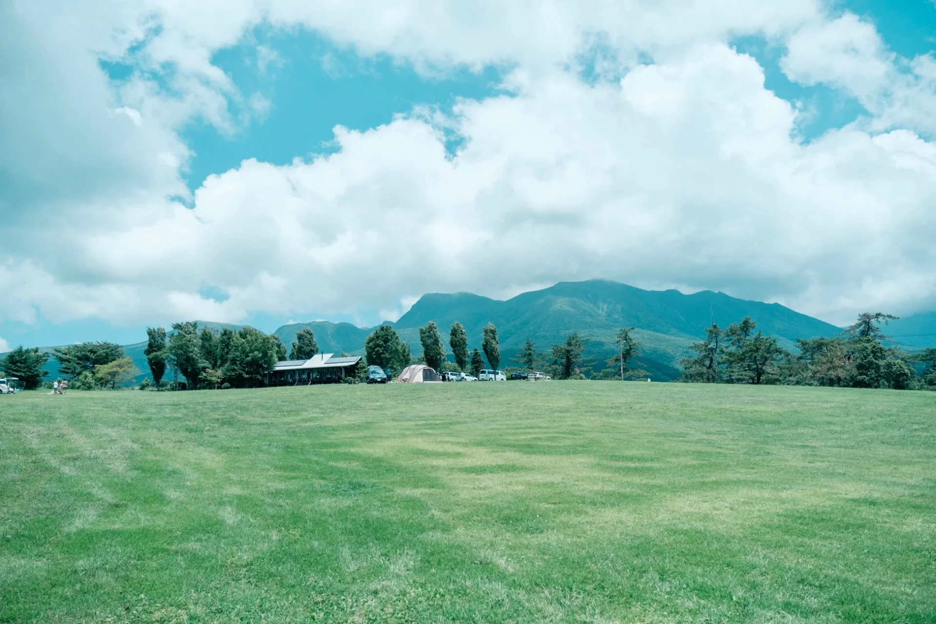 広々とした緑の草地が広がり、遠くには山々が見える風景です。青空には白い雲が浮かび、中景には建物、木々、駐車中の車、テントがあり、アウトドア施設やキャンプ場のような開放的な場所であることが伺えます。