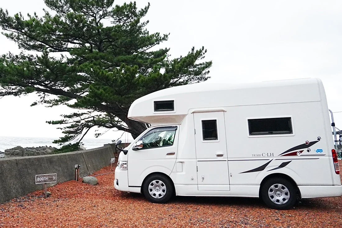 海辺の駐車場に白いキャンピングカーが停まっている風景