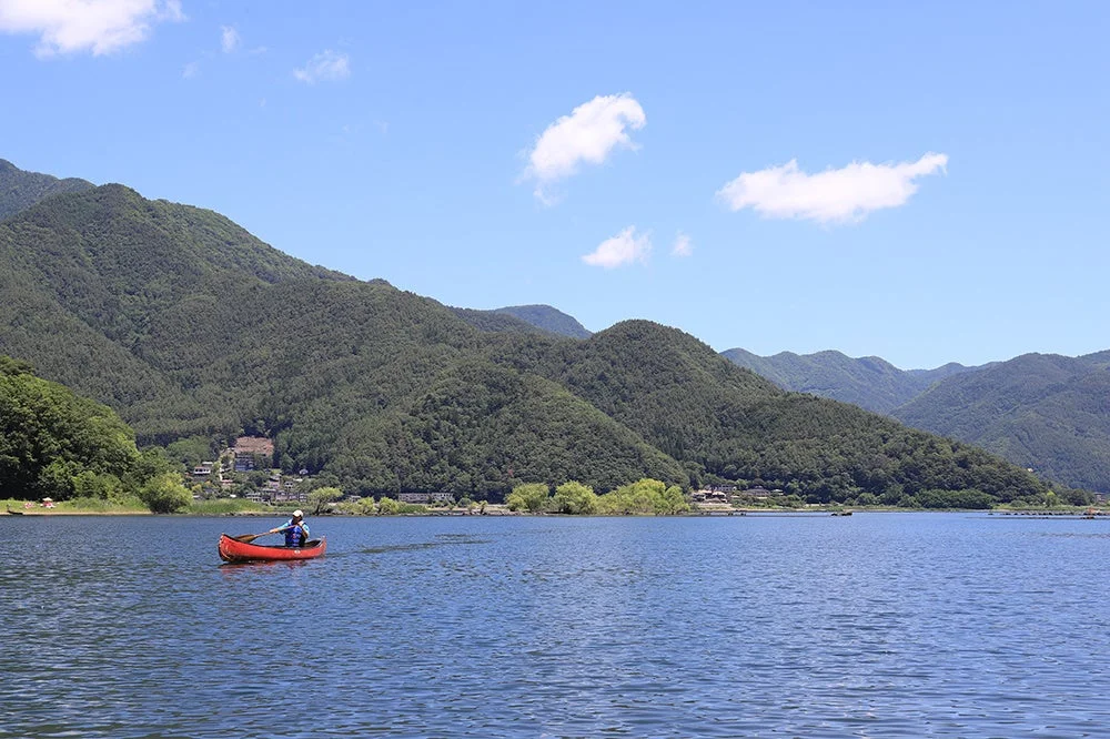 晴れた日、緑豊かな山々に囲まれた湖で、赤いカヌーに乗った人が穏やかに漕いでいる美しい風景です。青い空には白い雲が浮かび、水面は静かに輝いています。自然の中でのんびりとした時間が流れています。