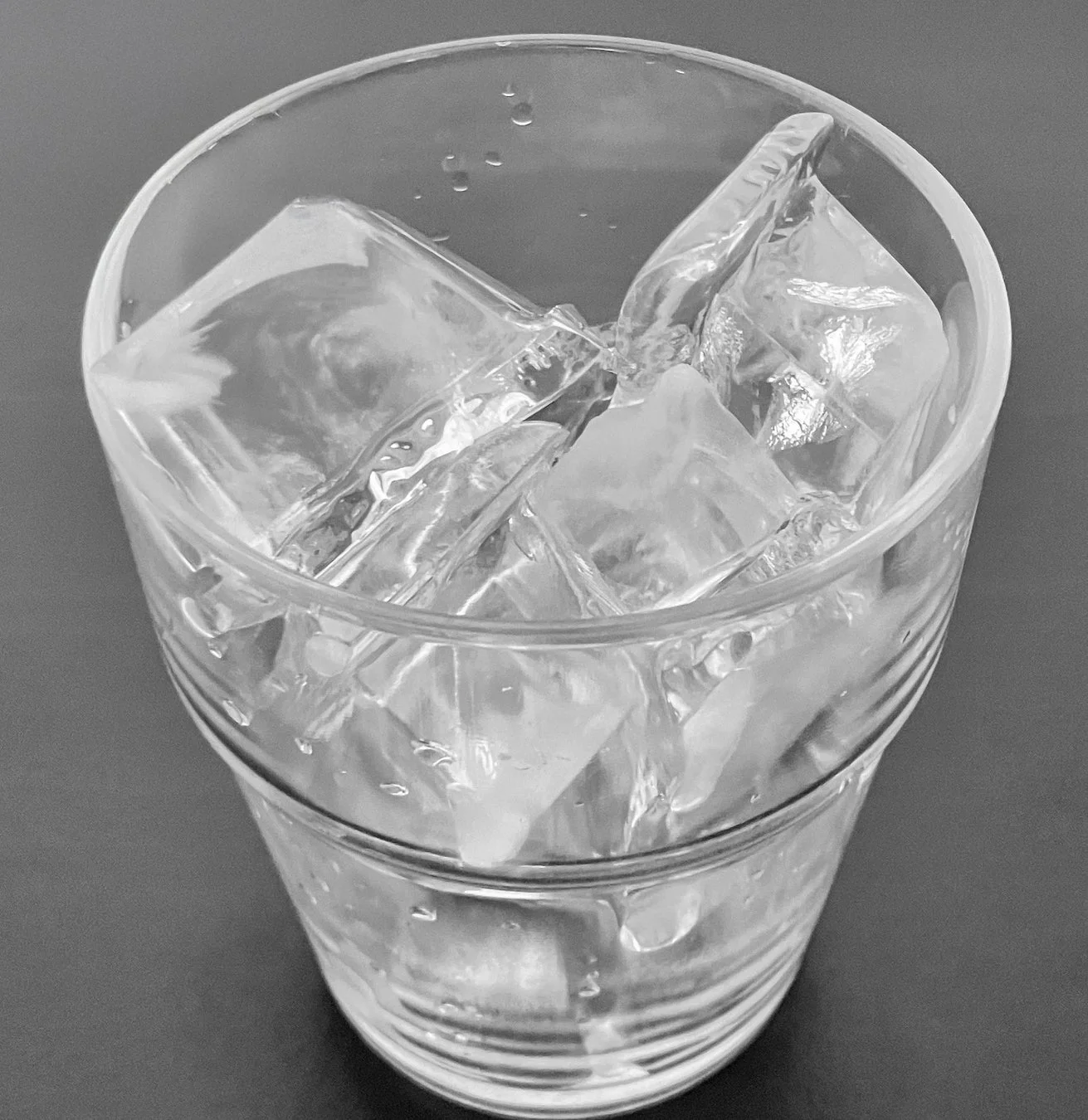 透明なグラスにたくさんの氷が入っている様子を上から捉えたモノクロ写真です。グラスの表面には水滴が見られ、冷たさが伝わってきます。