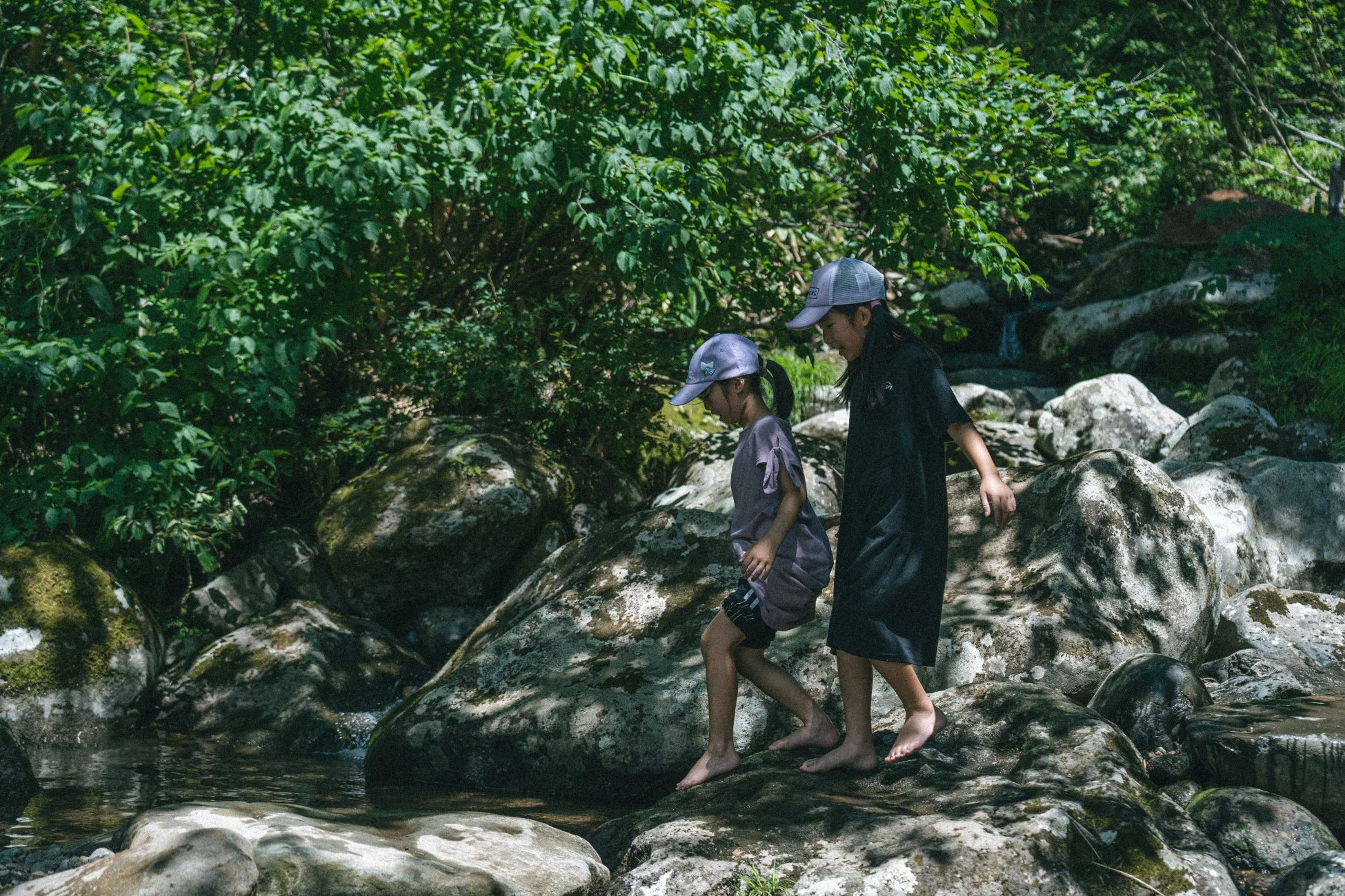 緑豊かな自然の中で、2人の子供が裸足で川の岩場を歩いている様子