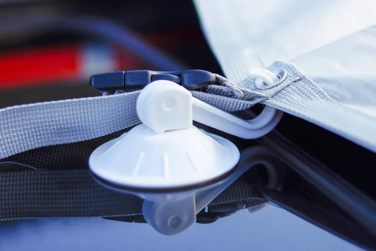 車のボディに固定された白い吸盤と、それに接続された灰色のストラップ、そしてシートの一部が写っています。防水シートやカーカバーなどを固定するためのアタッチメントです。