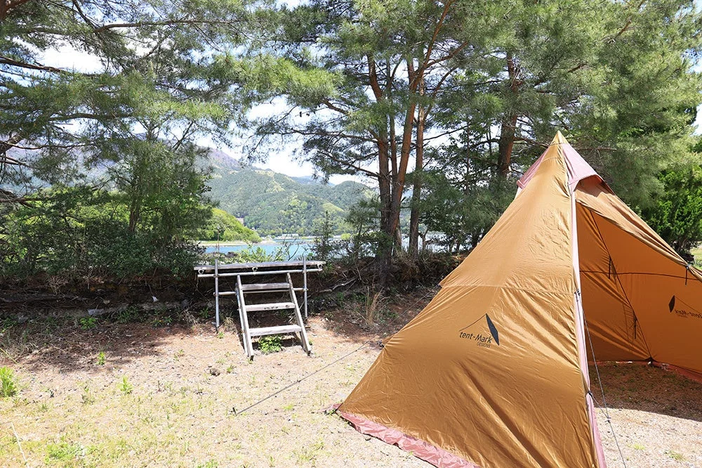 湖畔のキャンプ場で、茶色のテントが設営されています。背景には豊かな緑の山々と青い水面が広がり、自然を満喫できる穏やかなアウトドアシーンです。