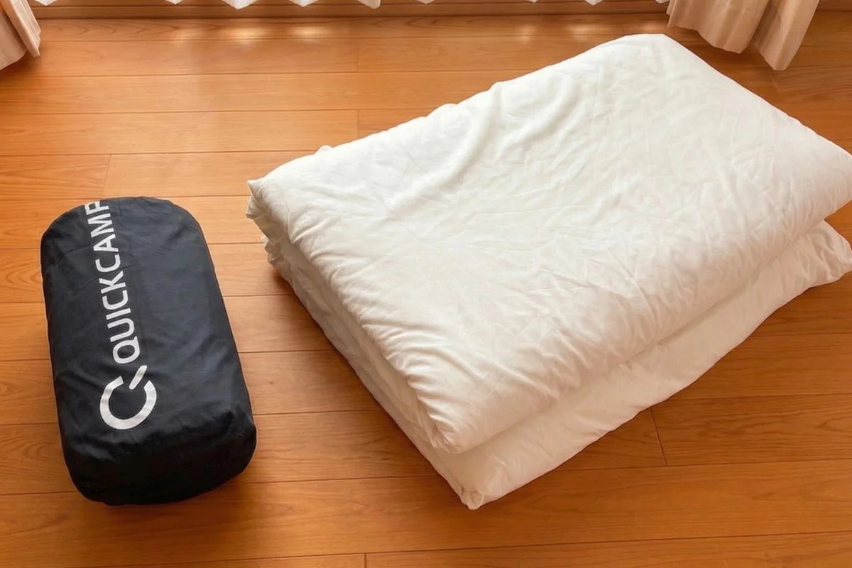 木のフローリングの上に、白い折り畳まれた布団とQUICKCAMPのロゴが入った黒い収納袋が並べられています