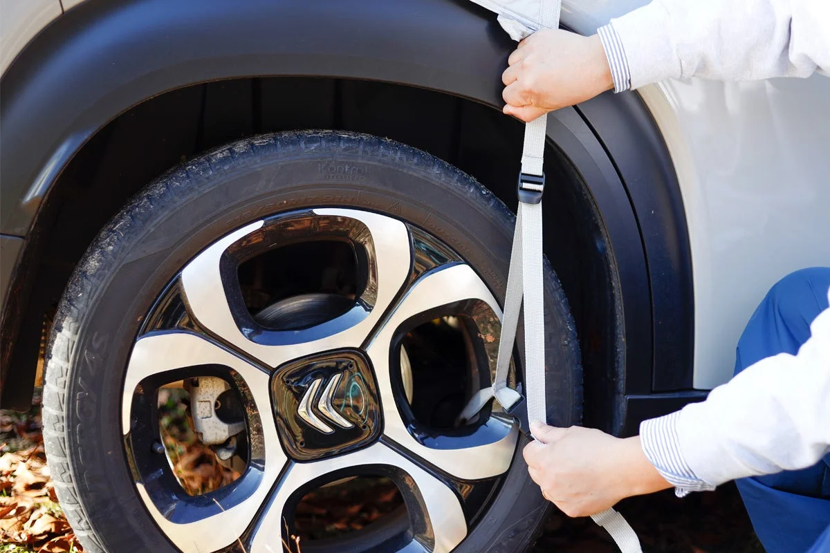 車のタイヤとホイールにストラップを取り付けている様子です。カーカバーを固定しているか、タイヤを保護する作業をしている手元がクローズアップされています。