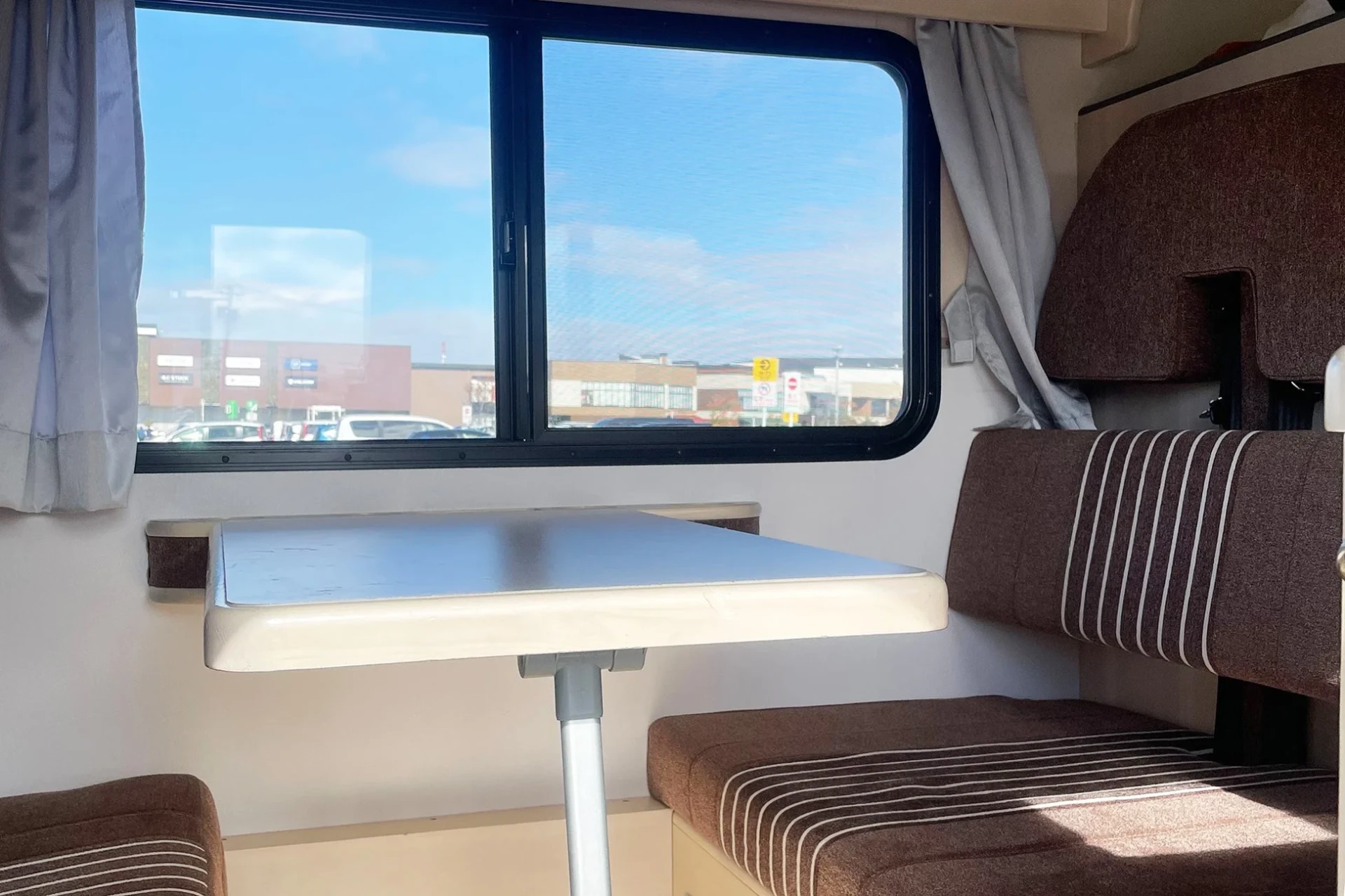 キャンピングカーの車内から外を眺めた様子。窓からは青空と駐車場、商業施設が見える