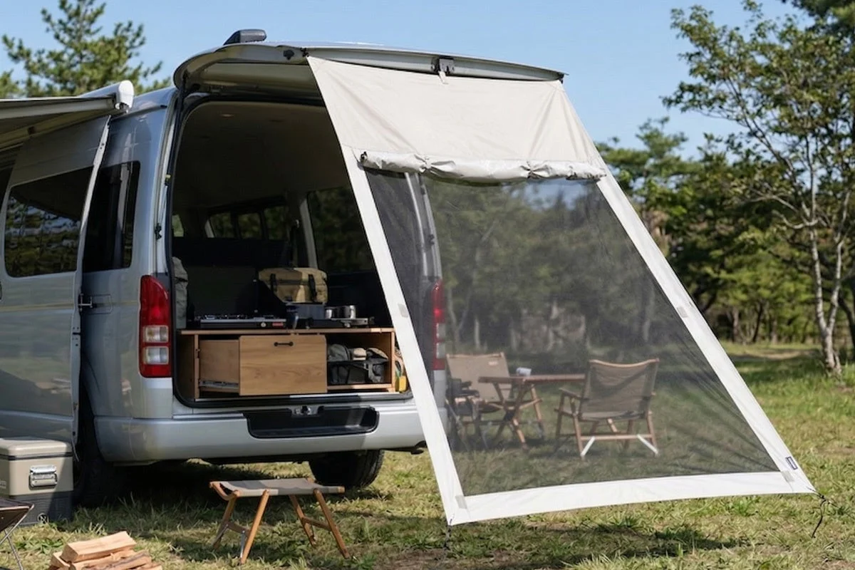シルバーのバンが自然の中でリアゲートを開け、蚊帳付きのタープを広げてキャンプを楽しんでいる様子。車内には調理器具が置かれ、屋外にはテーブルと椅子がセッティングされている。