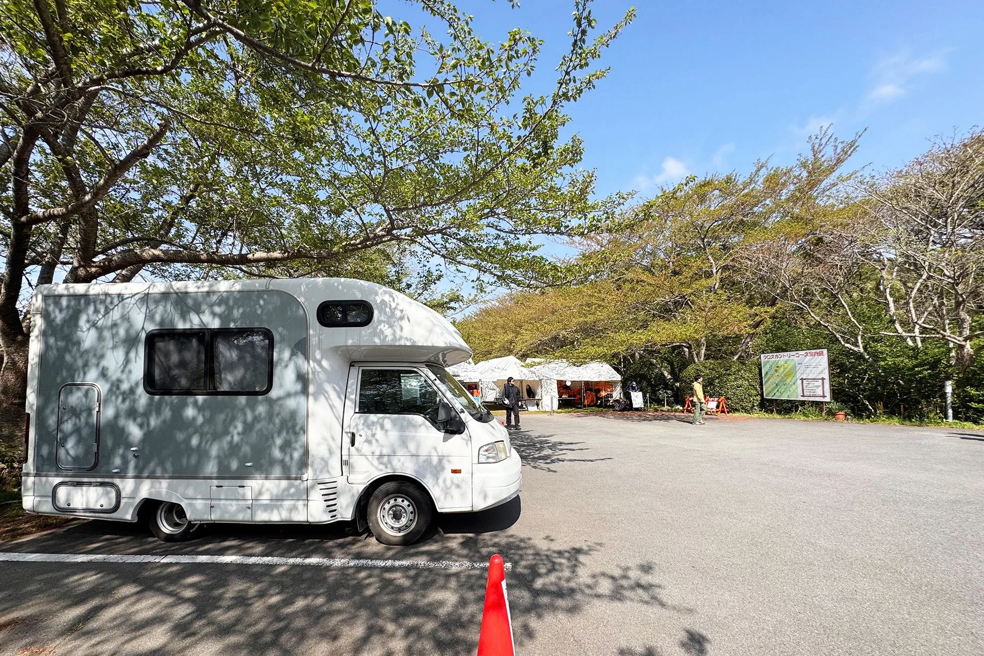 晴れた日にキャンピングカーが駐車され、背景に木々と白いテントが並ぶイベント会場