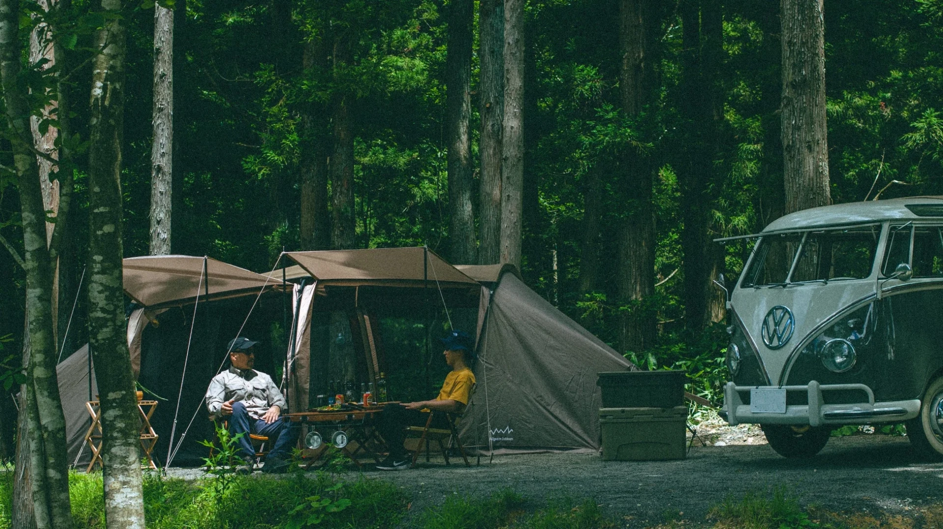 豊かな緑に囲まれた森の中で、ヴィンテージのフォルクスワーゲンバンとテントを設営し、二人がキャンプを楽しんでいる様子