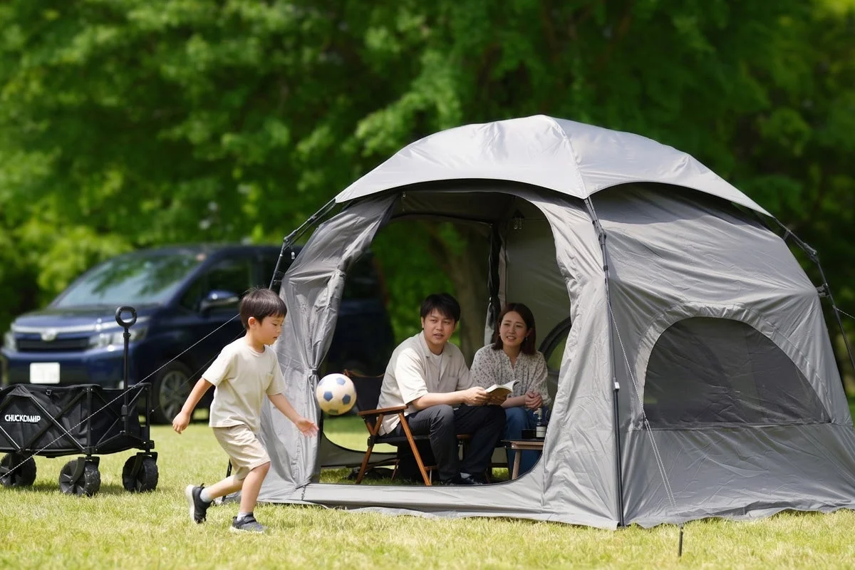 晴れた日に広々とした公園で、家族がアウトドアを楽しんでいます。息子がテントの外でサッカーボールを蹴り、両親はテントの中でリラックスして過ごしています。
