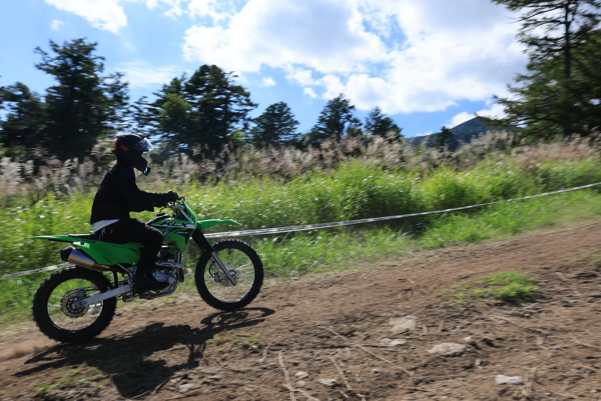 青空の下、緑色のオフロードバイクに乗ったライダーが土のコースを走行している様子を捉えた写真です。背景には草木が生い茂り、遠くに山が見えます。