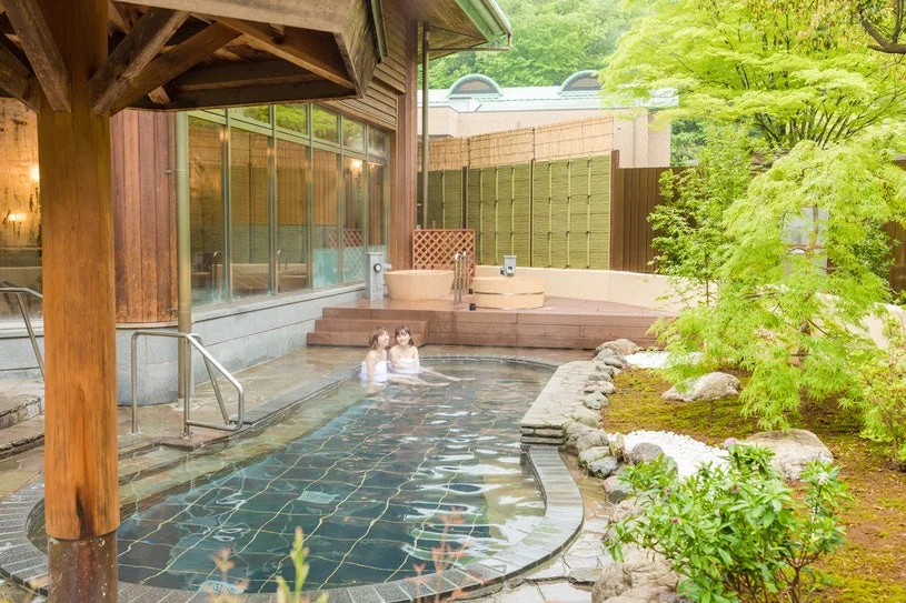 日本の旅館の露天風呂で、新緑に囲まれた庭園を眺めながら二人の女性がくつろいでいる様子。和風の建物と自然が調和した癒しの空間です。