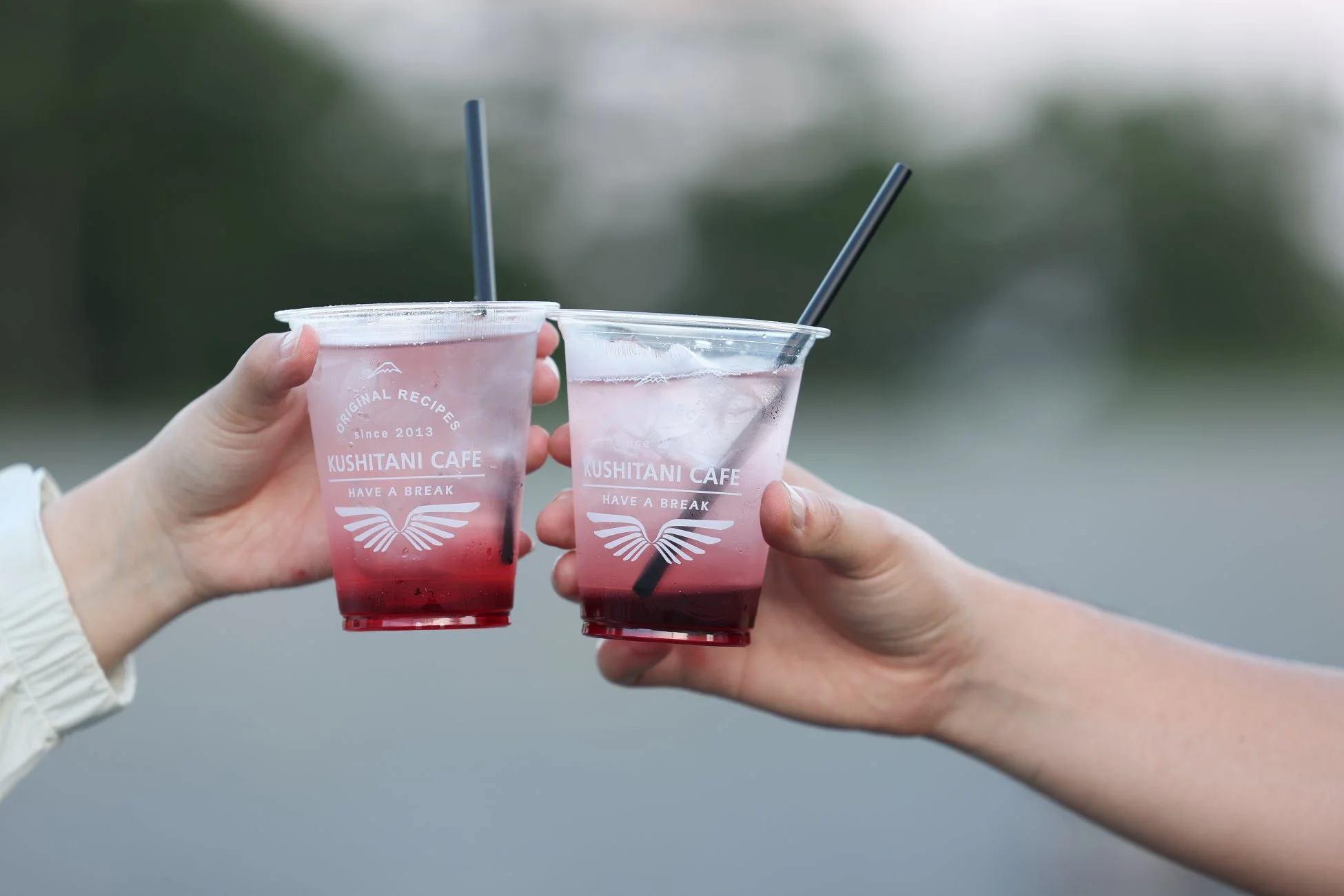 「クシタニカフェ」のロゴが入ったクリアカップに入った赤いグラデーションのドリンクを、二人がストローで持っている様子。リフレッシュや休憩のひとときを捉えた写真です。
