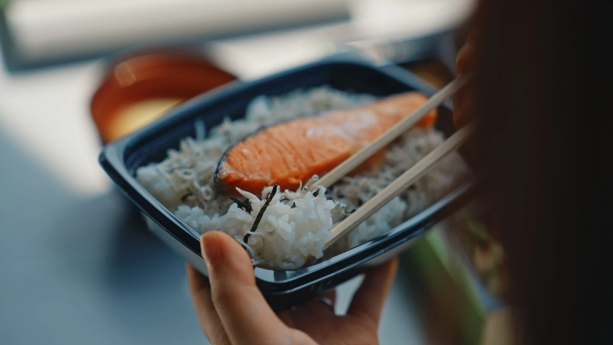 鮭とご飯、しらすが入った弁当を箸でつまんでいる様子を写した画像です。手軽な和食ランチの風景です。