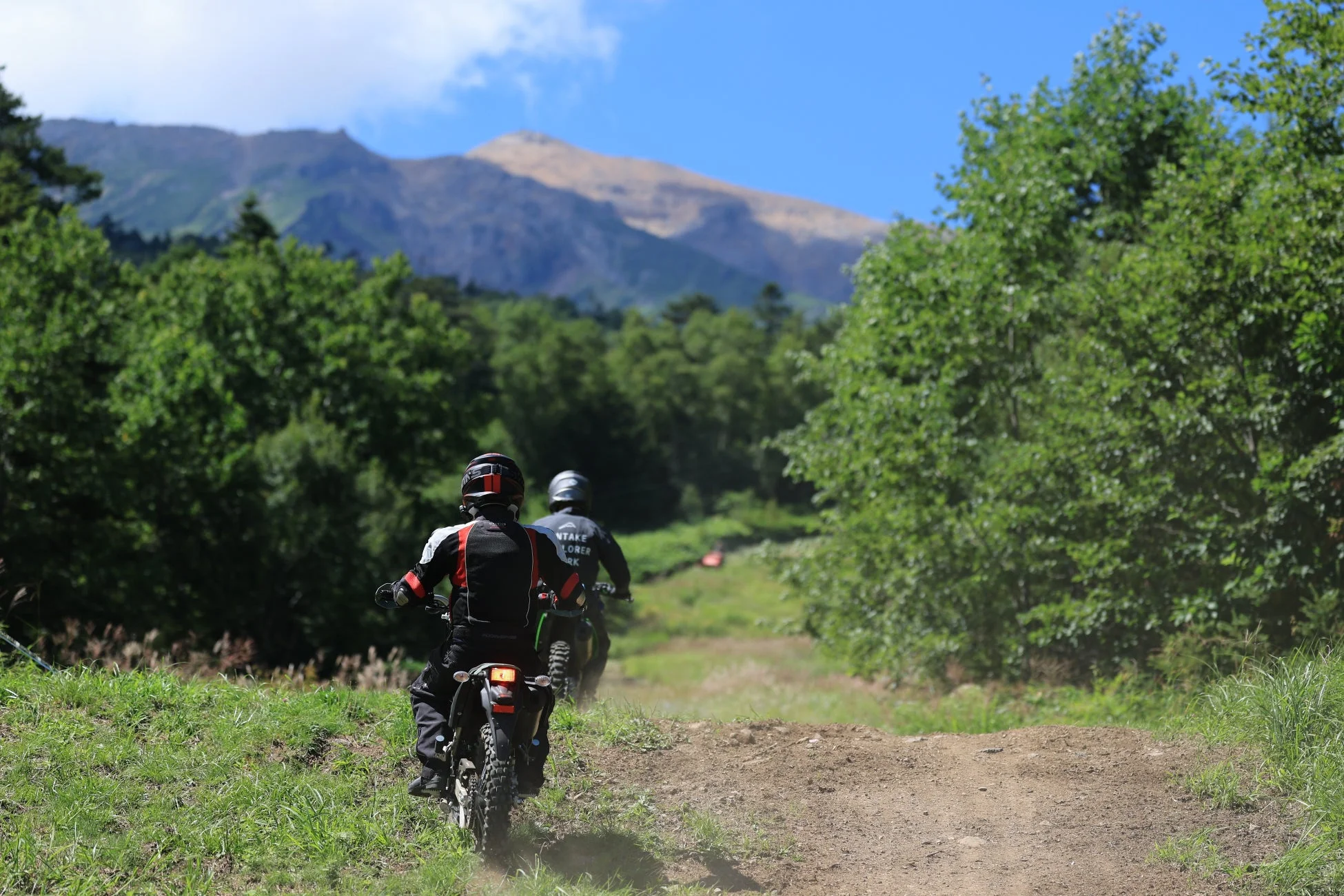 青空の下、緑豊かな山間部の未舗装路をオフロードバイクに乗った2人のライダーが走行している様子です。遠くには大きな山が見え、自然の中でのツーリングを楽しんでいるように見えます。