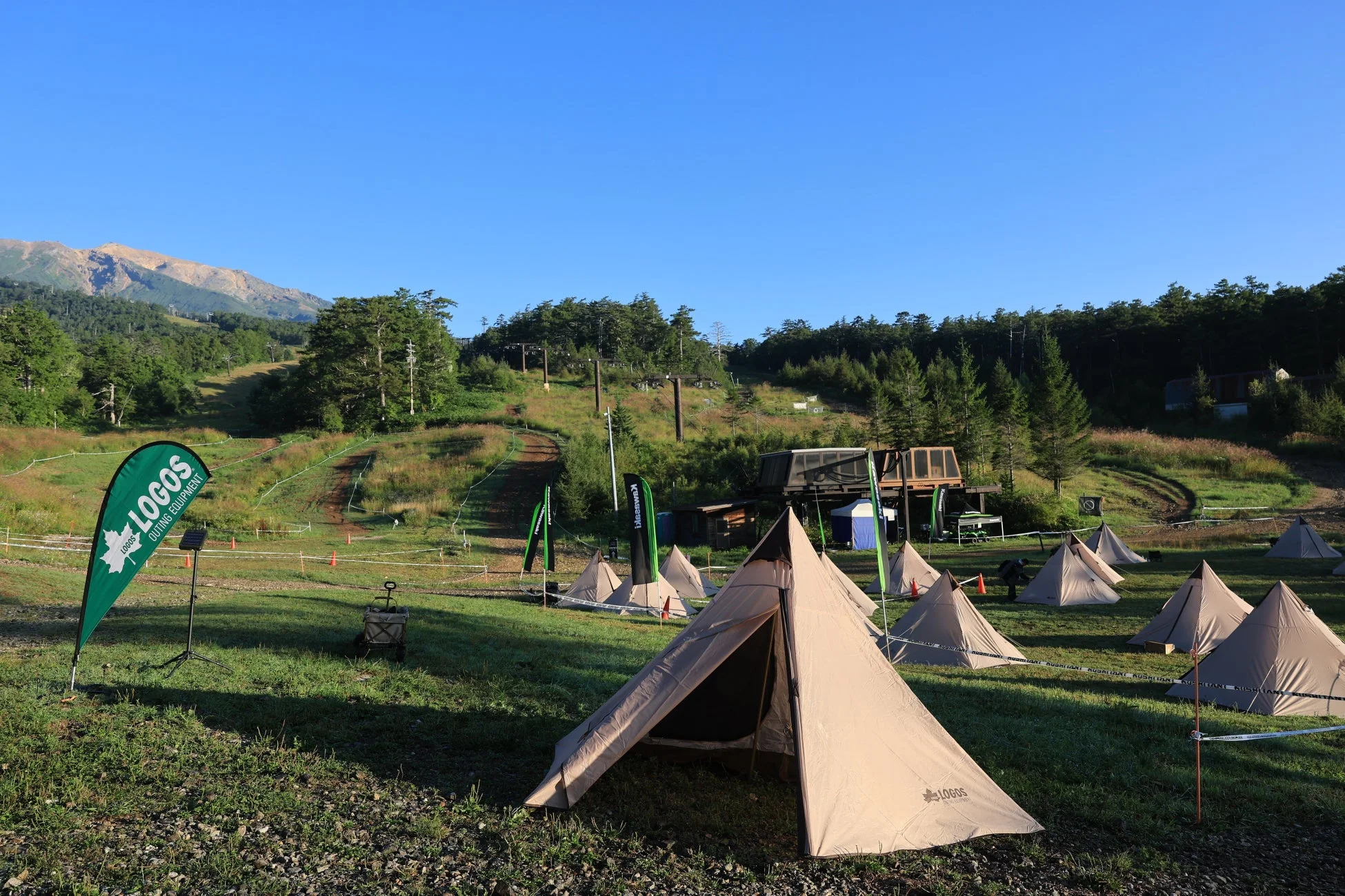 夏の晴れた日、山間の広々としたゲレンデ跡地で多数のテントが設営されているアウトドアイベントの様子。ロゴスやクシタニ、カワサキの旗が見られます。