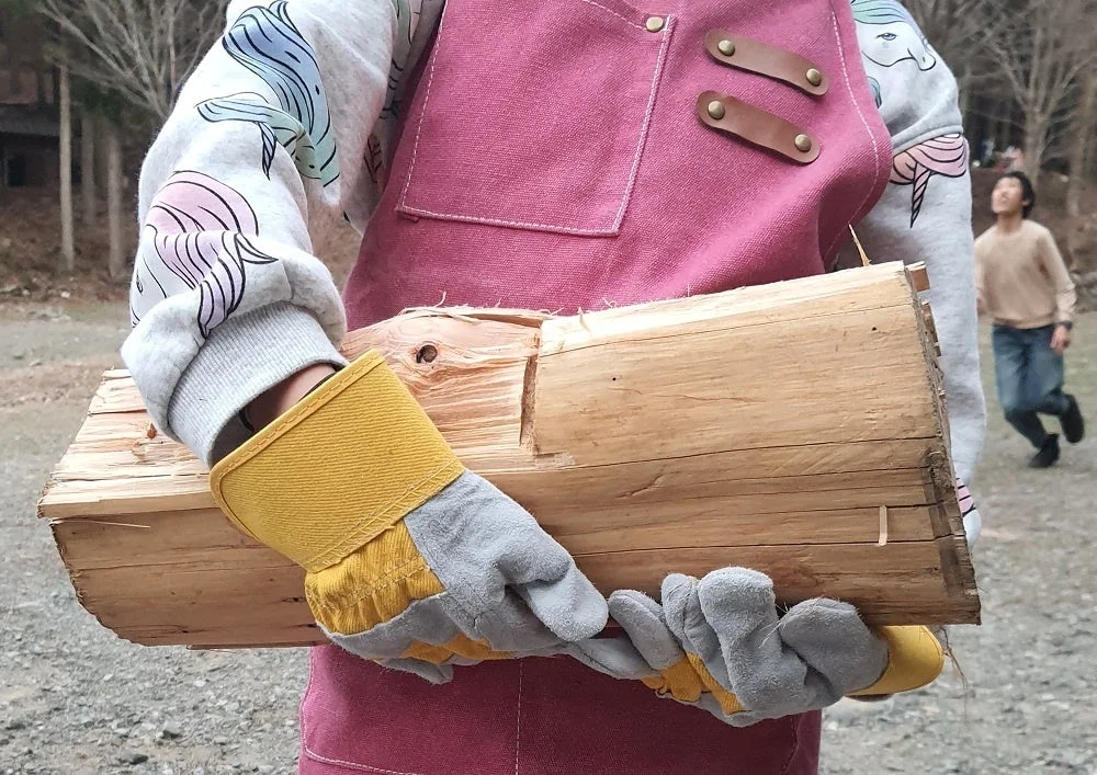 手袋とエプロンを着用した人物が、割られた薪を抱えて運んでいます。