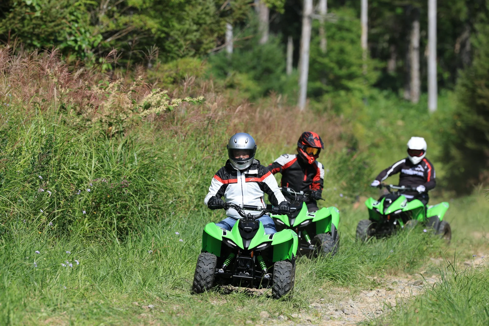 3人のライダーが緑色のクワッドバイクに乗り、ヘルメットを着用して森の中のオフロードコースを走行しています。アウトドアでのアクティビティを楽しんでいる様子が伺えます。