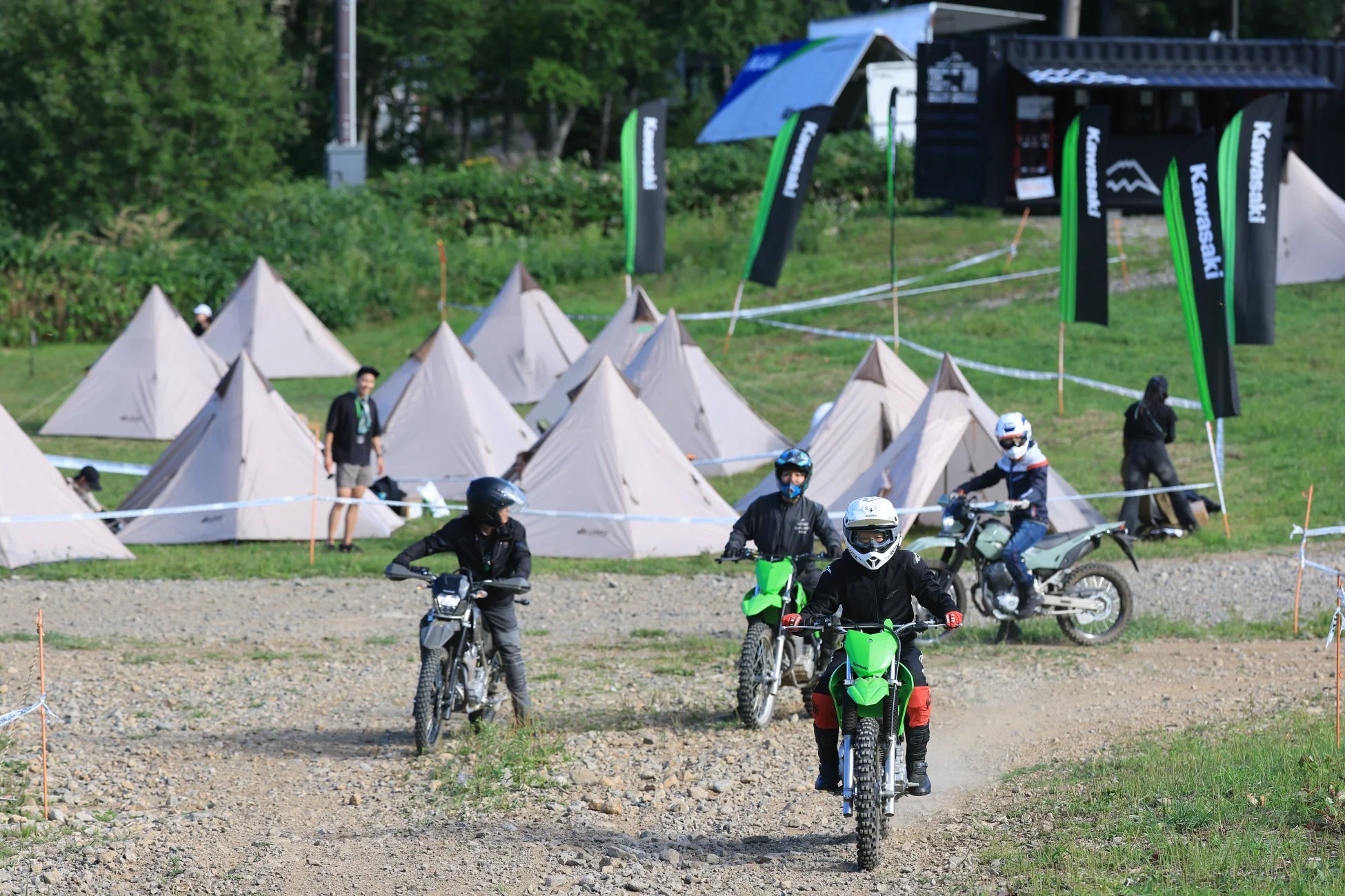 河原のような場所でオフロードバイクに乗る人々が映っています。背景には多数のテントが並び、Kawasakiの旗が複数立っていることから、バイクイベントやキャンプイベントの様子を捉えたものと思われます。