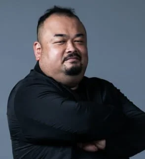 黒いシャツを着たぽっちゃり体型の男性が、腕を組み、笑顔で目を細めているポートレート写真です