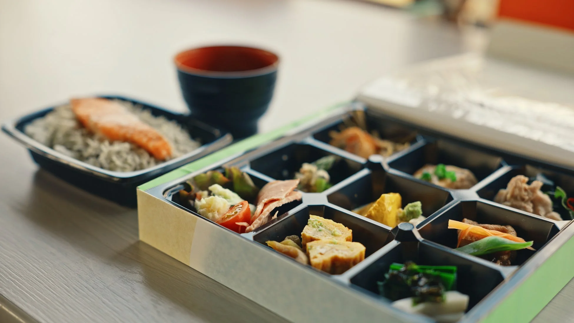 様々な種類のおかずが詰められた豪華な弁当箱と、鮭が乗ったご飯が写っています。手前には味噌汁椀も見え、和食の食事が準備されている様子です。