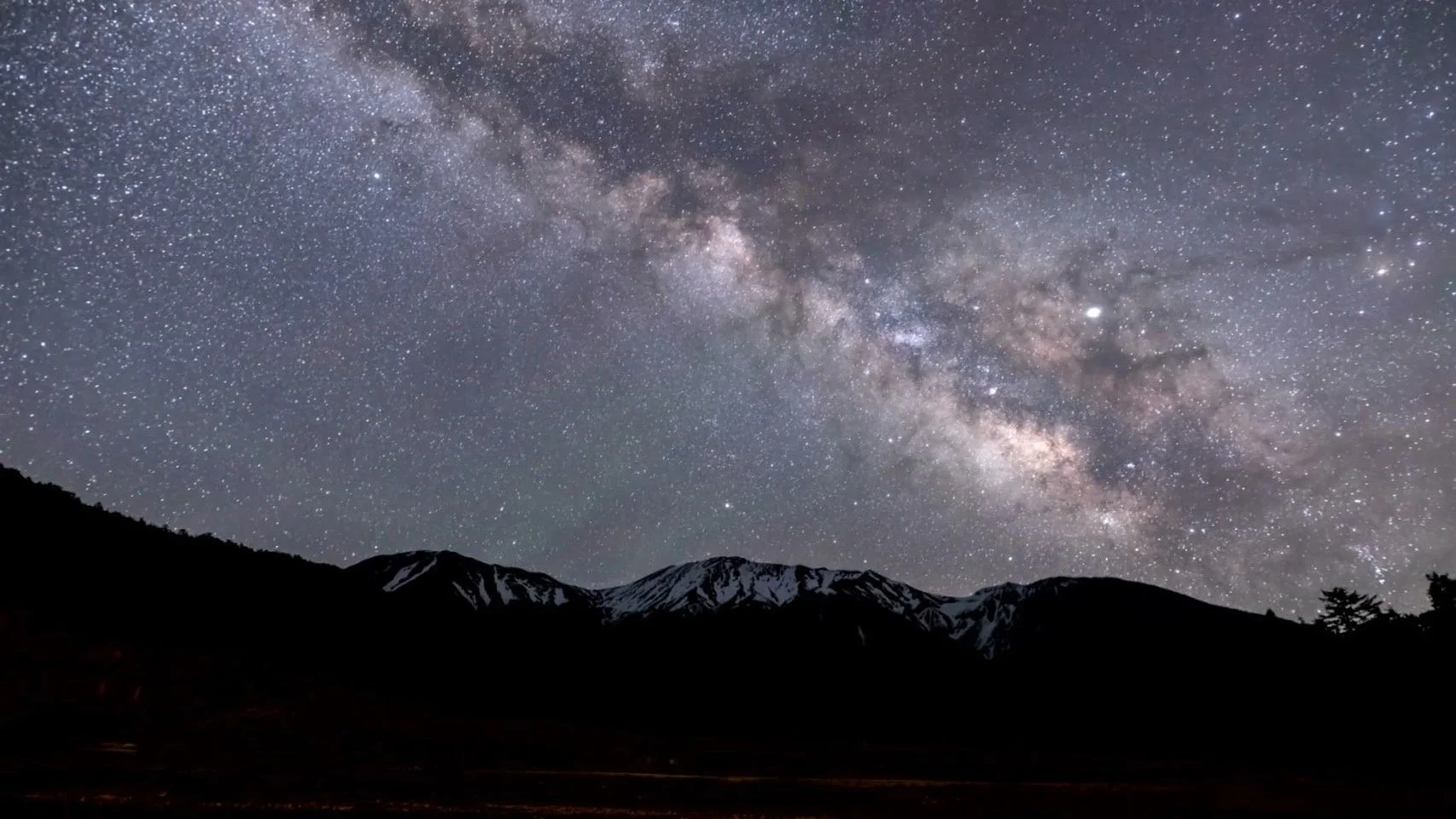 雪を抱いた山々のシルエットの上に、無数の星々が輝く壮大な天の川が広がる夜景です。