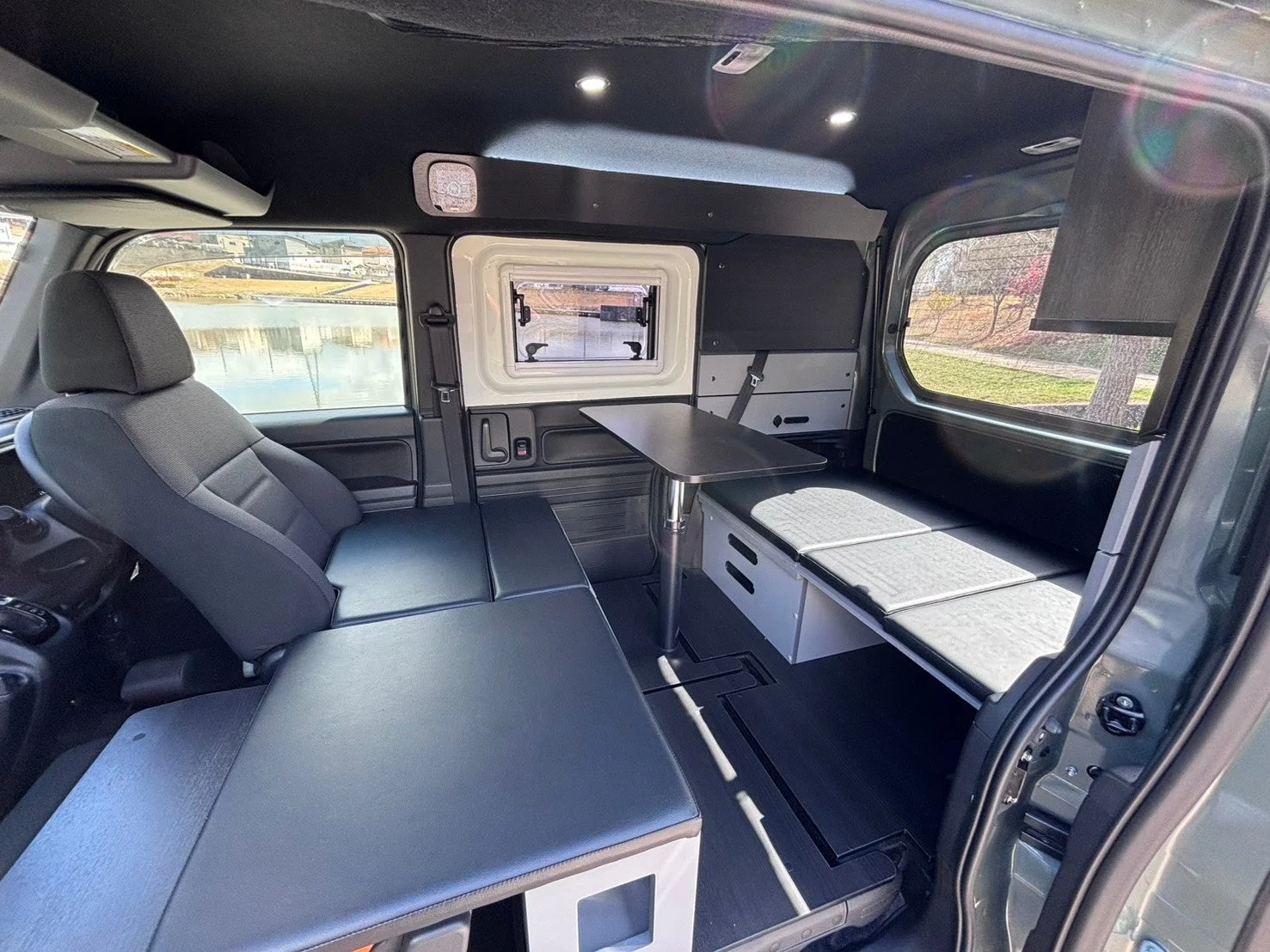 キャンピングカーの車内を写した画像で、運転席の後ろにベッドとして使用できる座席があり、中央には折りたたみ式のテーブルが設置されています