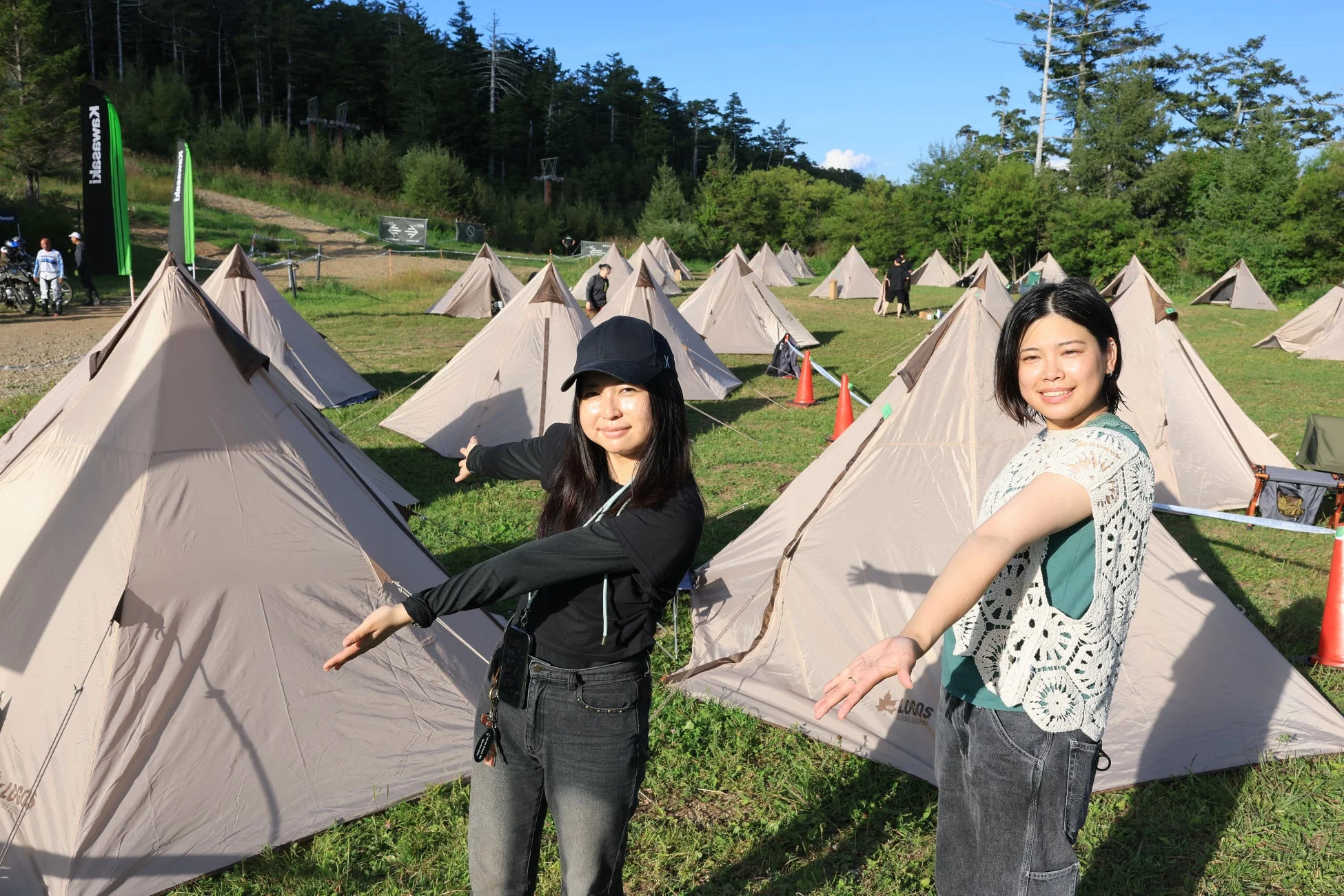 晴れた屋外の広場で、笑顔の2人の女性が多くのテントを背景にポーズをとっています。キャンプイベントかアウトドアの集まりのようです。