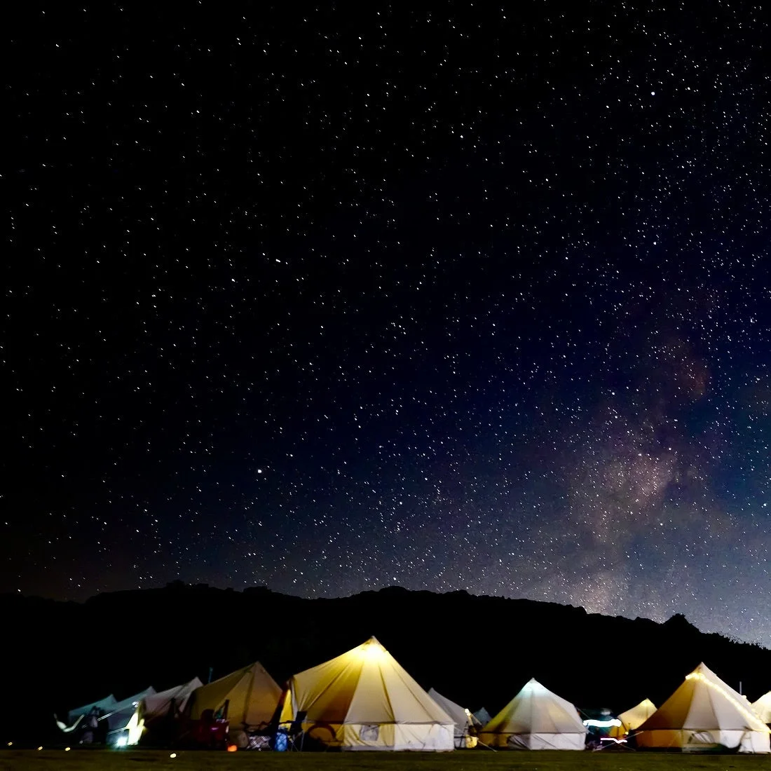 満天の星が輝く夜空の下、山々を背景に複数のベルテントが並ぶキャンプ場