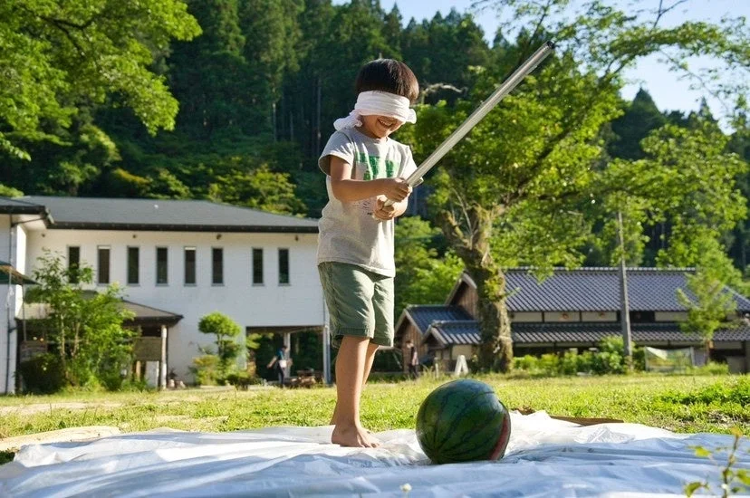 目隠しをした子供が屋外でスイカ割りをしている様子です。笑顔で棒を構え、夏の伝統的な遊びを楽しんでいる瞬間が捉えられています。背景には豊かな緑と建物が見えます。