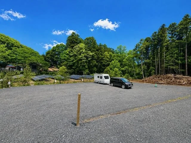 晴れた青空の下、広々とした砂利の駐車場にキャンピングカーと黒い車が停まっています。周囲には緑豊かな森が広がり、一部に太陽光パネルも設置されている自然豊かな風景です。
