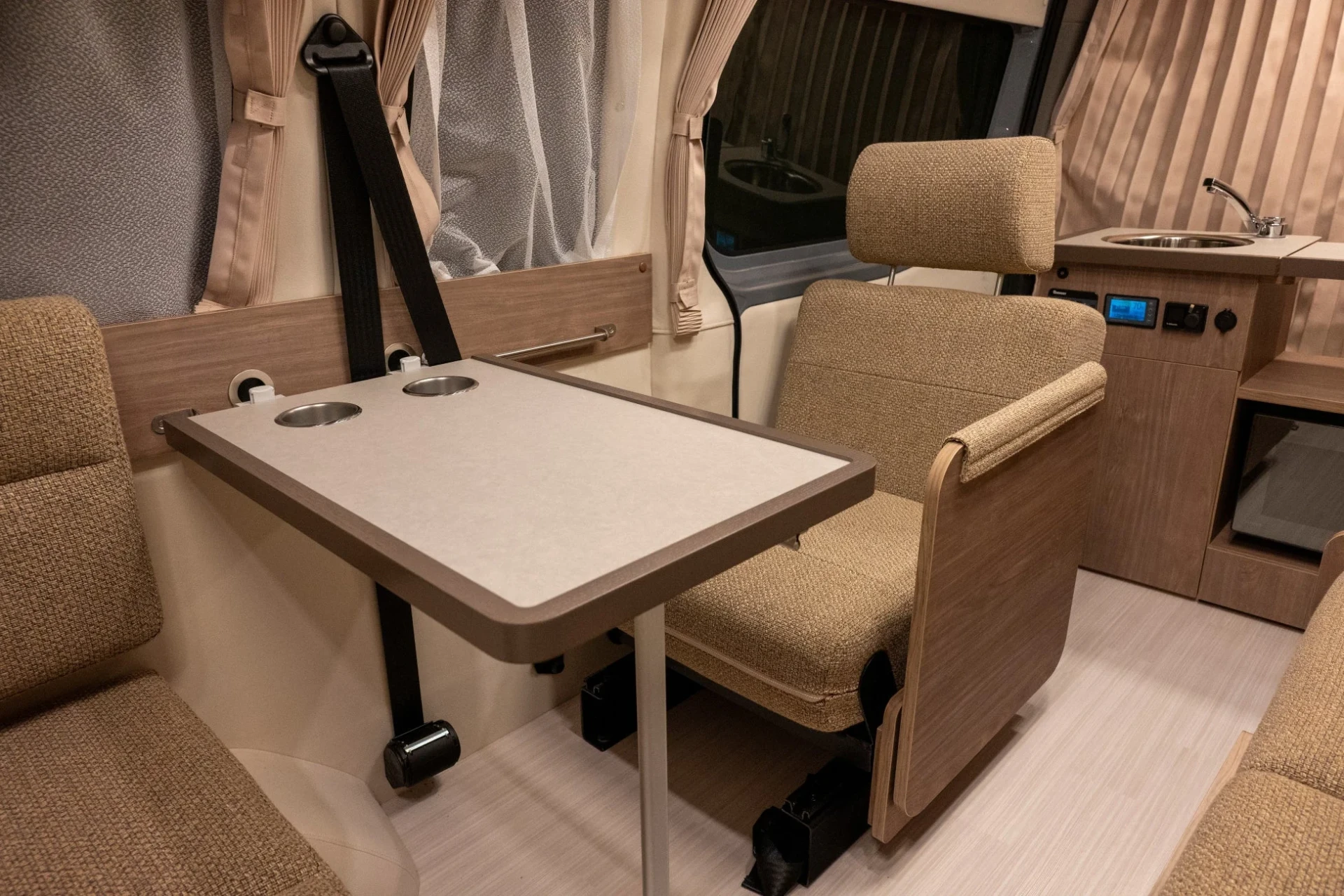 モダンなキャンピングカーの車内インテリア。ベージュ系のシートとテーブルが配置され、コンパクトなキッチンシンクも備わった、機能的な居住空間が広がっています。