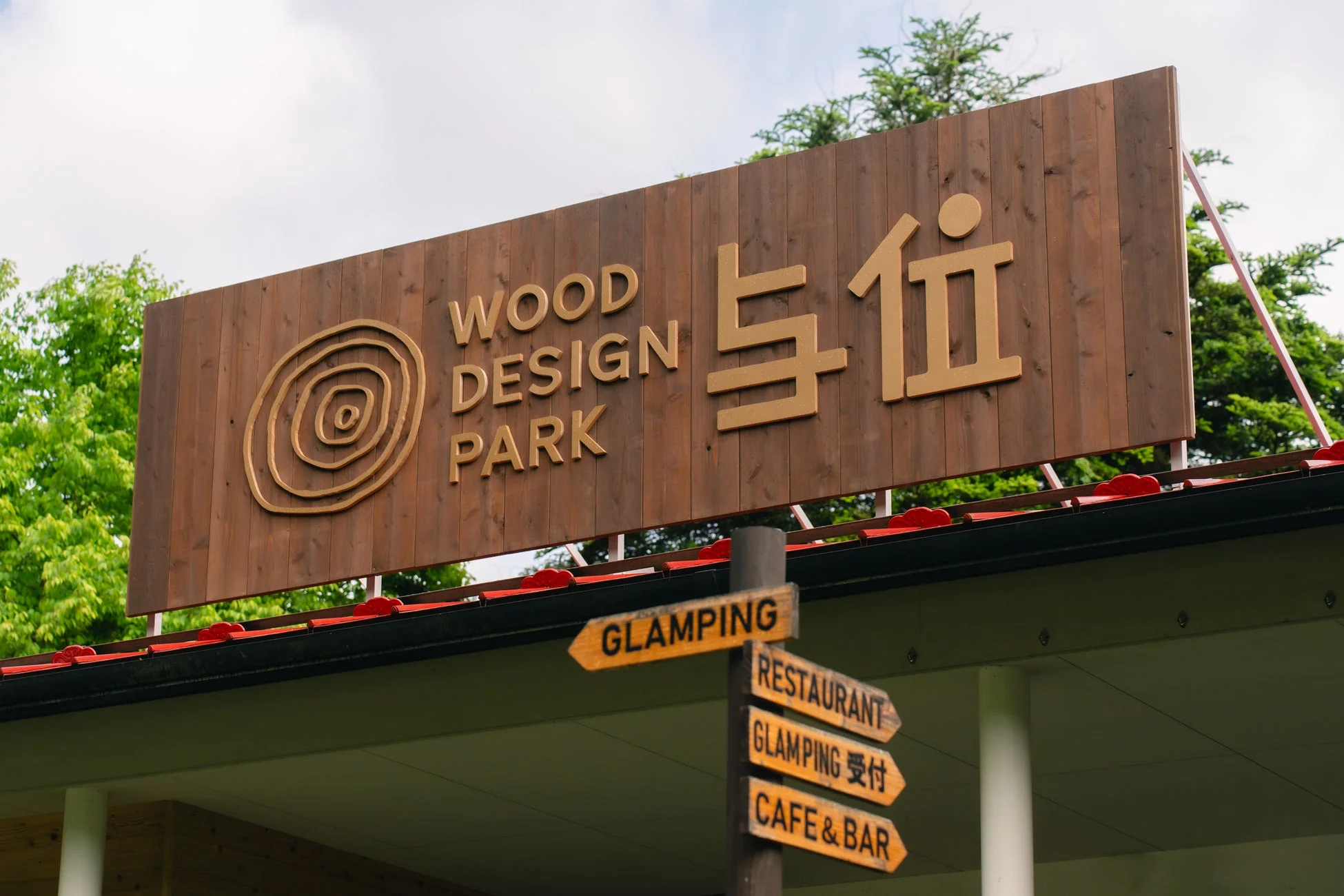 「WOOD DESIGN PARK 与位」の木製看板と、グランピング、レストラン、カフェ＆バーへの方向を示す案内板が写っています。自然の中にあるレジャー施設の入り口付近の様子です。