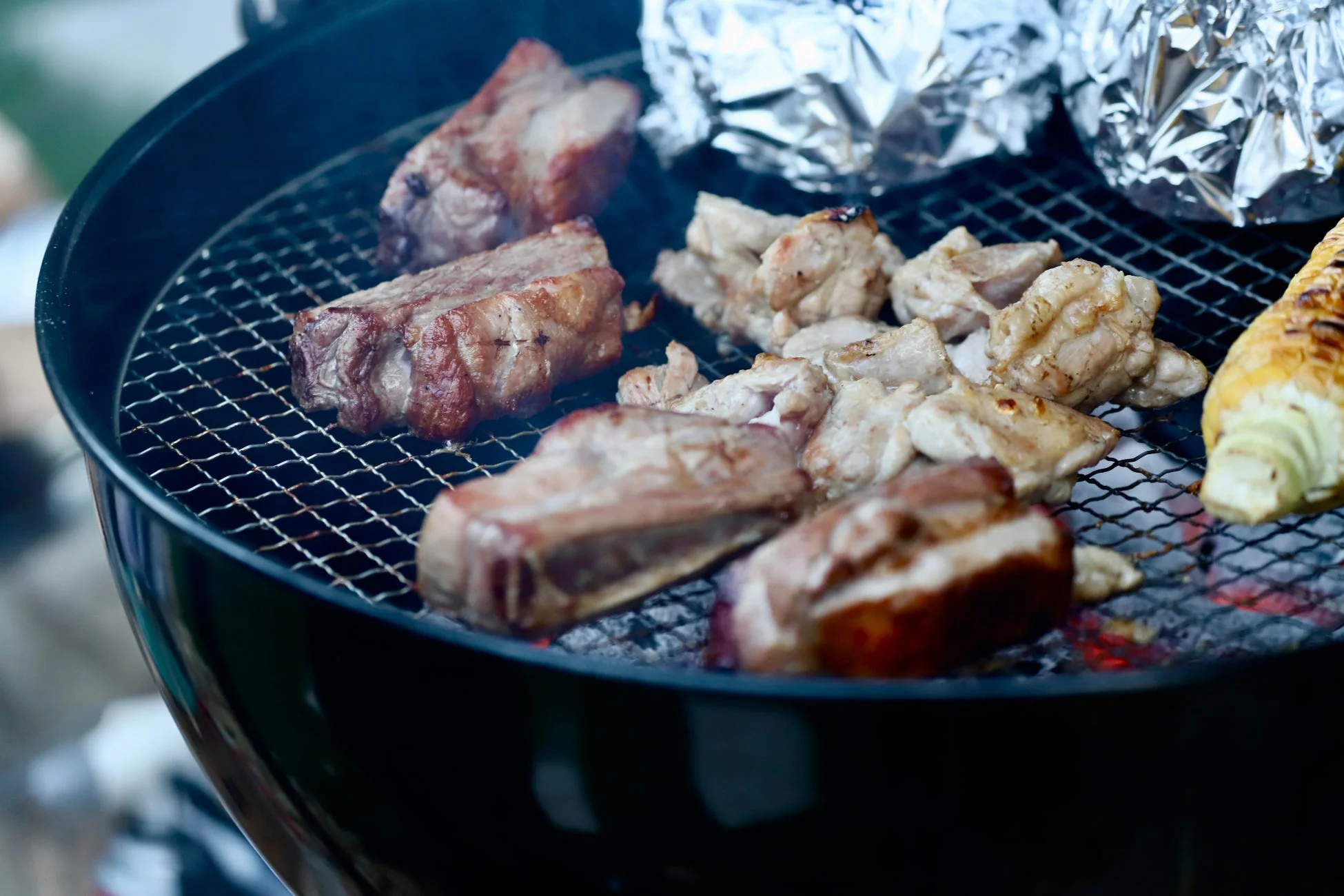 バーベキューグリルで、骨付き肉や鶏肉、とうもろこしなどが炭火で焼かれている様子を捉えた画像です。アルミホイルに包まれた食材も見え、食欲をそそるアウトドア料理の風景が広がっています。