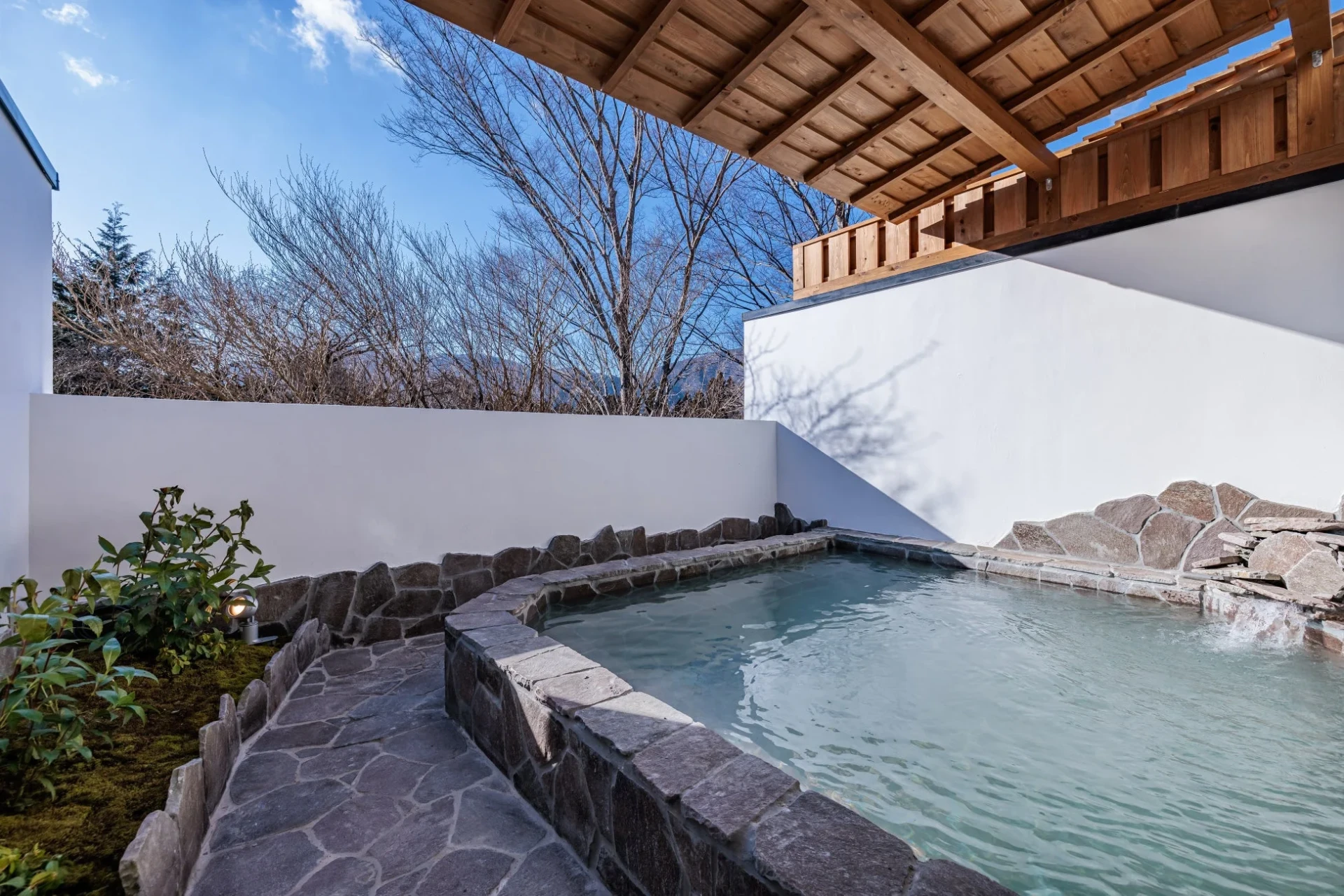 冬の晴れた日に撮影された、石造りの露天風呂の風景。白い壁と木の屋根に囲まれ、湯気が立つ温泉と、背景には葉を落とした木々が広がっています。和風で落ち着いた雰囲気が漂う、リラックスできる空間です。