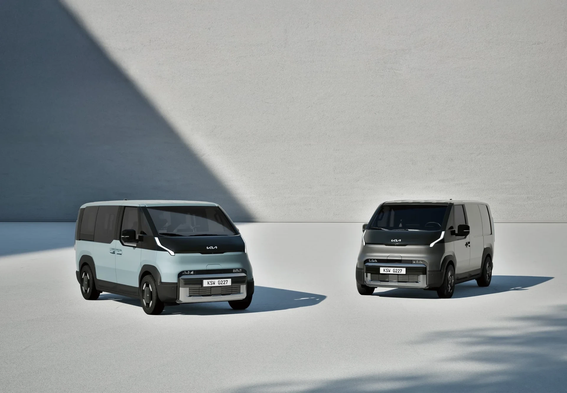 現代的なデザインのKiaブランドのバンが2台、シンプルな背景に並んで駐車されています。水色と濃い灰色の車両が、未来的な印象を与えています。