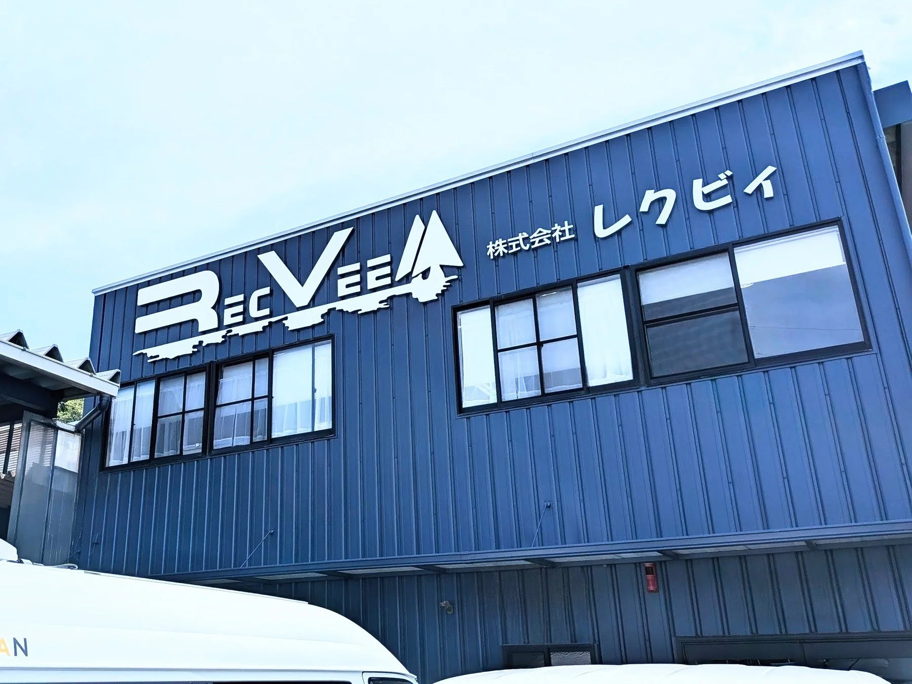 青い波板の外壁を持つ会社の建物が写っています。白い文字で「RECVEE 株式会社 レクビイ」と書かれた看板が特徴的です。窓がいくつか並び、建物の手前には白い車両の一部が見えます。