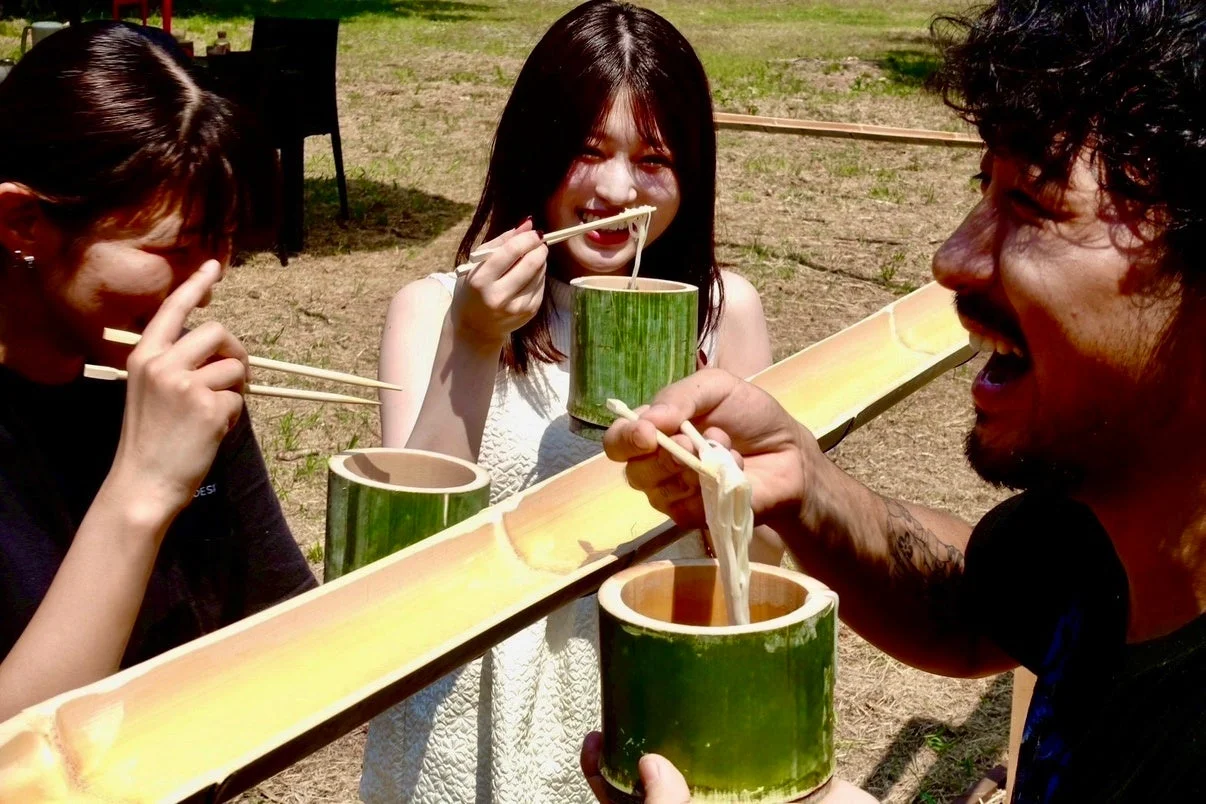 夏の屋外で、3人の男女が流しそうめんを楽しんでいます。笑顔で箸を使い、竹の樋から流れてくるそうめんをすくっており、日本の夏の風物詩を満喫している楽しげな光景です。