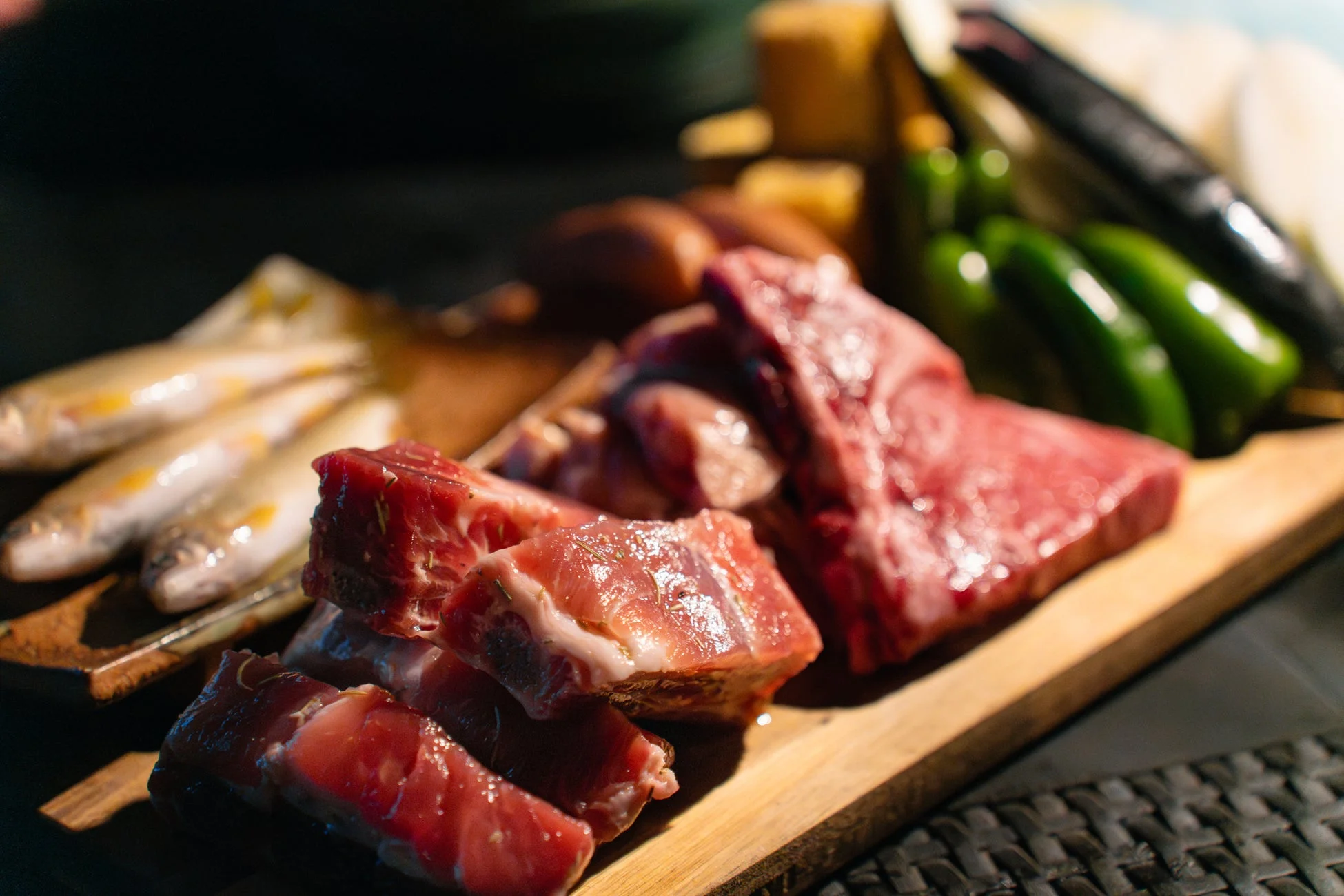まな板の上に新鮮な生肉（ステーキ肉と骨付き肉）、小魚、ピーマン、ナスなどの食材が並べられています。バーベキューやグリル料理の準備段階であることが伺える、食欲をそそる一枚です。