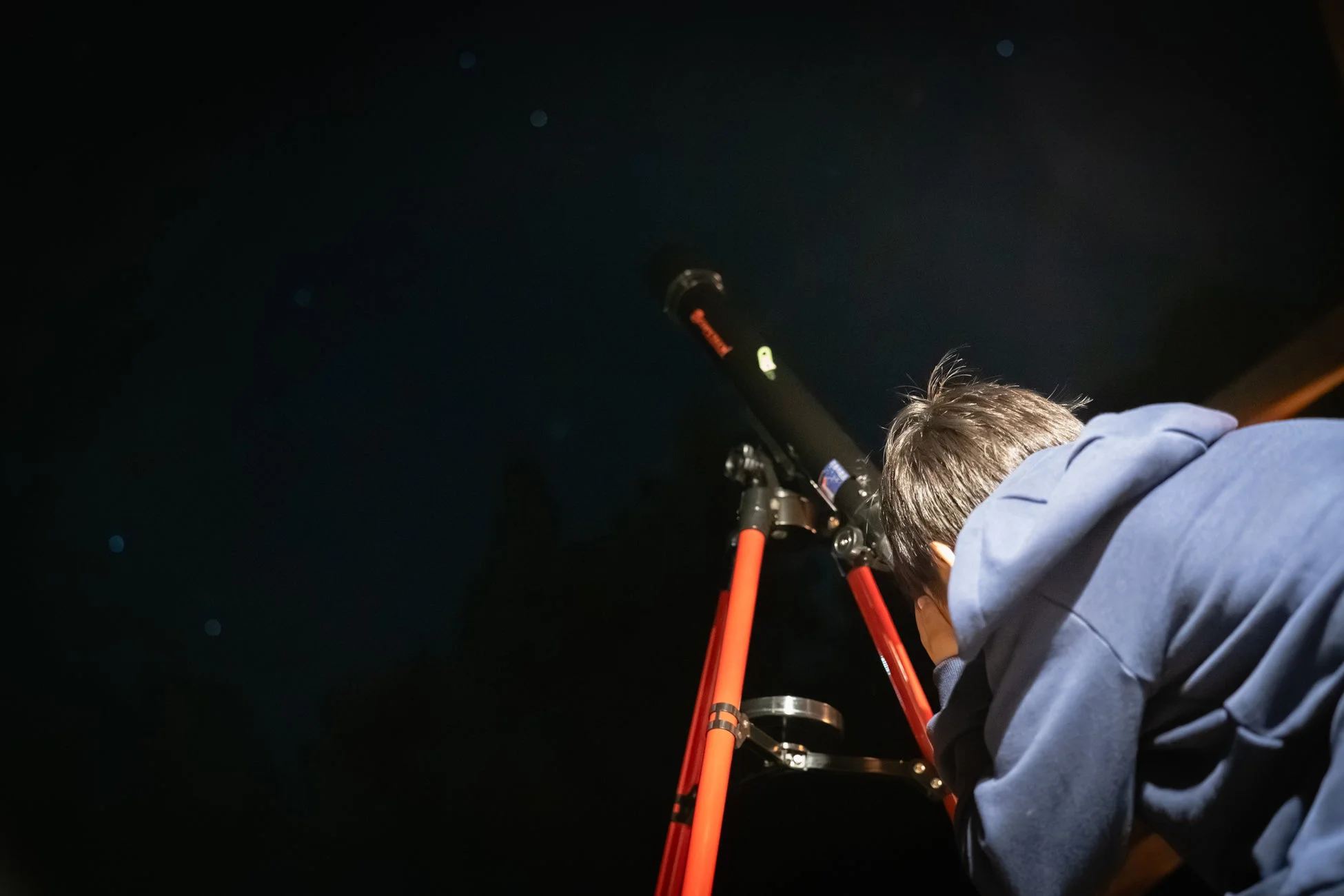 暗い夜空の下、一人の人物が望遠鏡を覗き込み、星や天体を観測している様子です。天体観測の瞬間を捉えた写真です。
