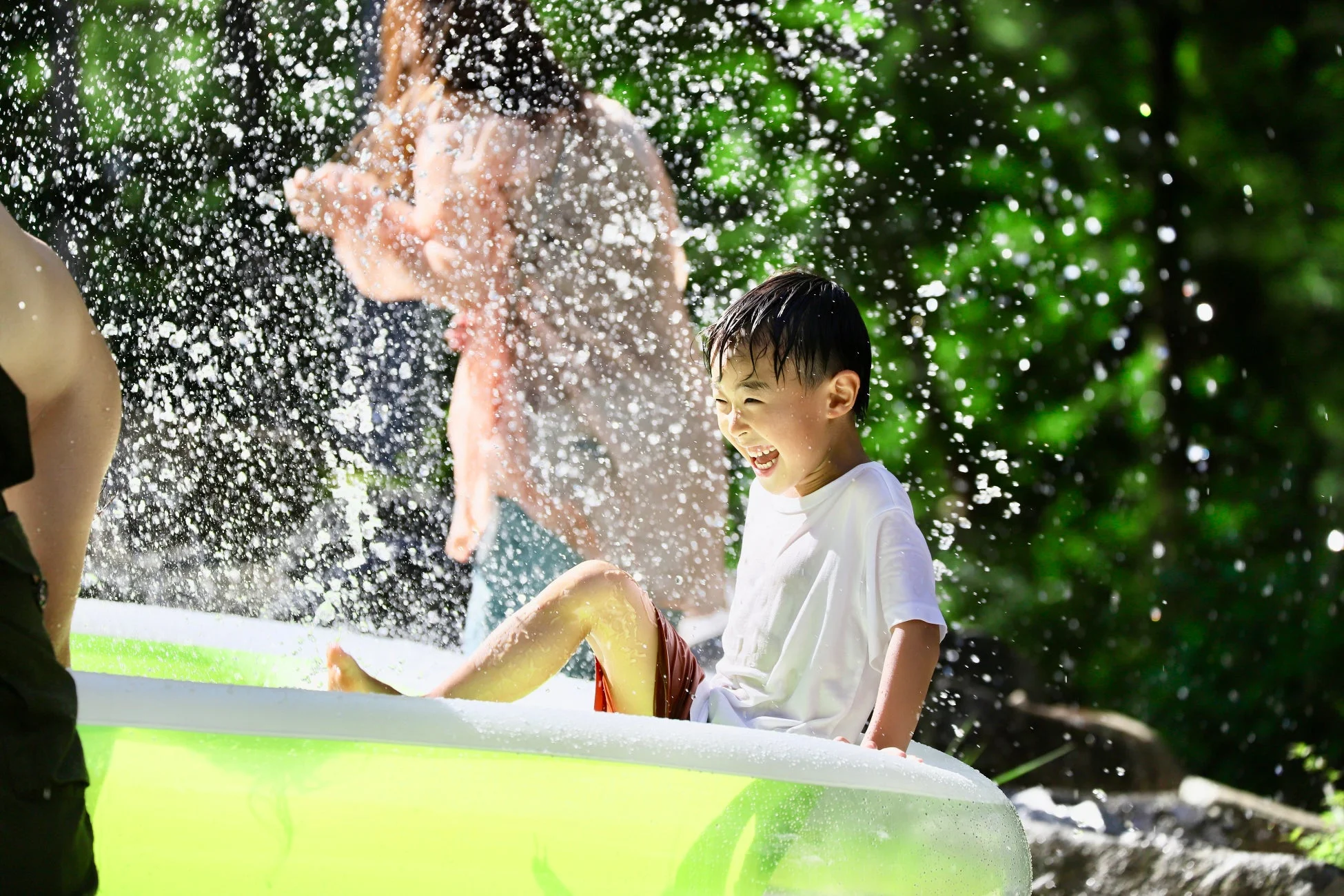 屋外で子供がビニールプールに入り、水しぶきの中で満面の笑みを浮かべて楽しんでいる様子。夏の水遊びの楽しさが伝わる一枚です。