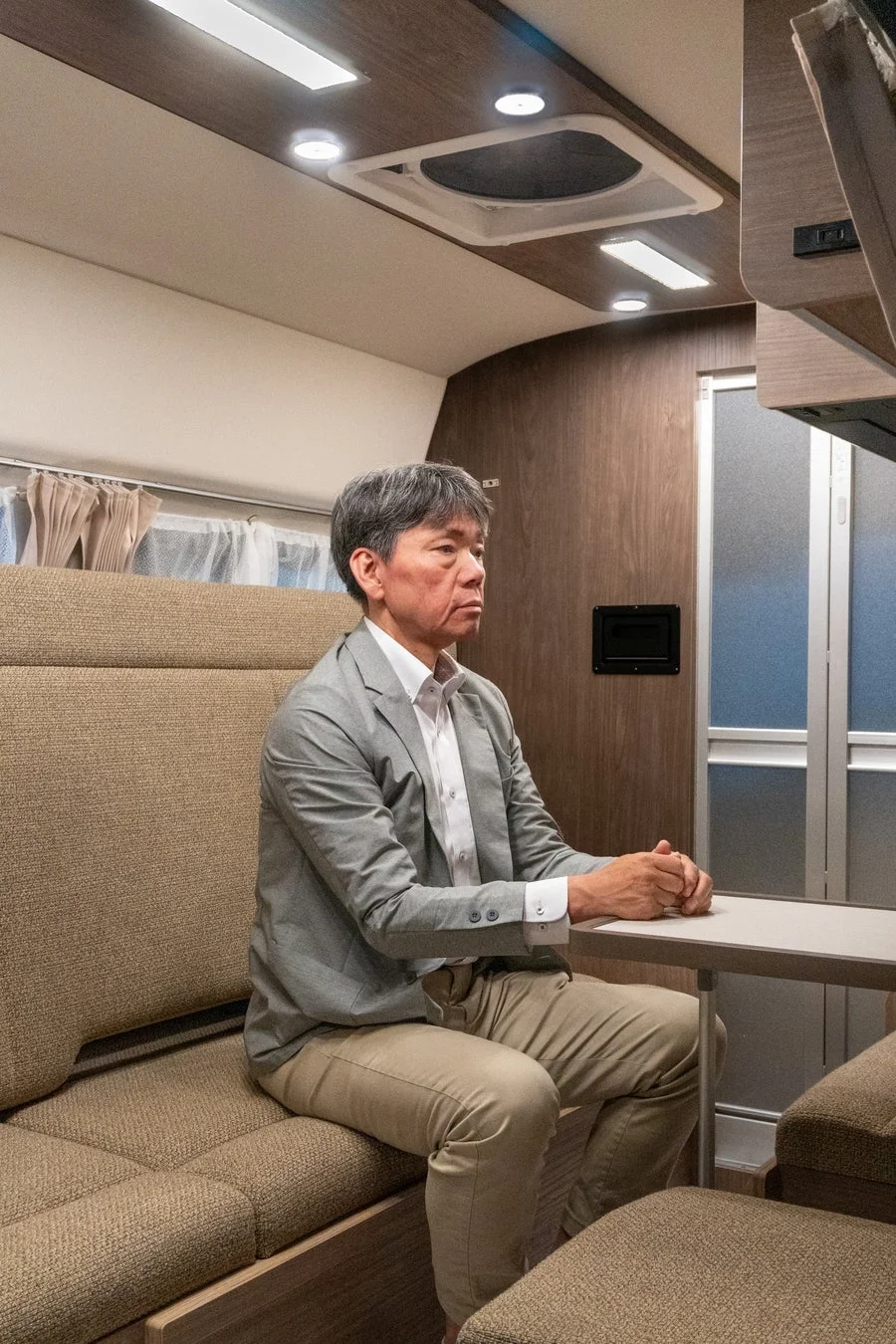 キャンピングカーのような車内で、スーツ姿の男性がテーブルに手を組み座っている。木目調の壁と明るい照明が特徴の落ち着いた空間。