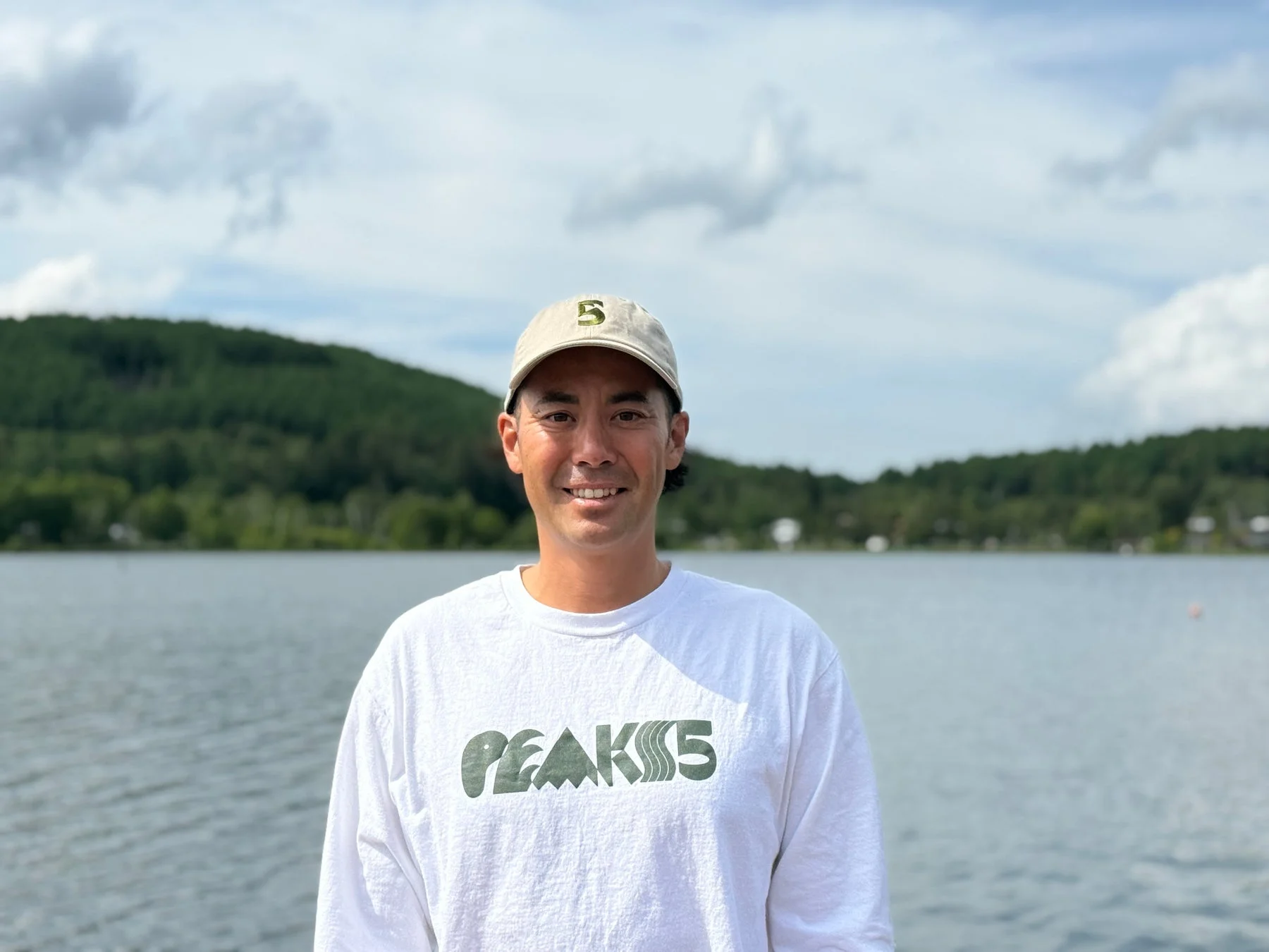 湖と緑豊かな山々を背景に、白い長袖Tシャツと帽子を着用した男性が笑顔で立っているポートレート