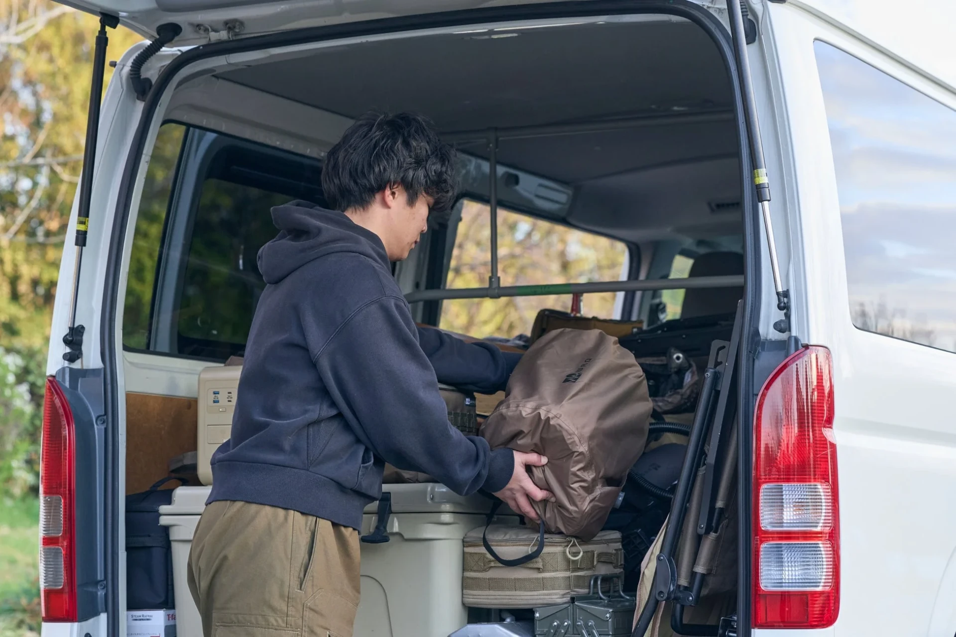 白いバンの開いたトランクで、男性がアウトドア用品やキャンプギアを整理し、積み込んでいる