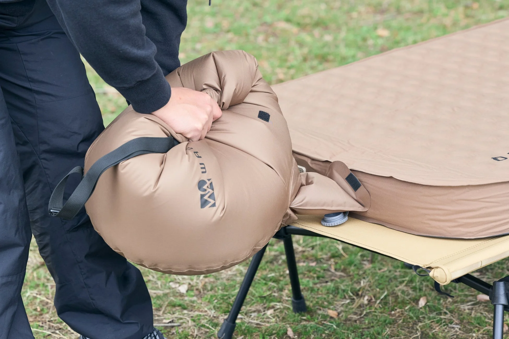 屋外で、人がキャンプ用コットの上にインフレーターマットを広げ、空気を入れているか、抜いている様子