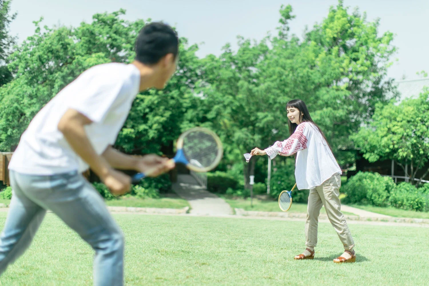 晴れた日に公園の芝生でバドミントンを楽しむ男女の様子。女性は笑顔でシャトルコックを構え、男性はラケットを持って対峙しています。緑豊かな背景が爽やかな雰囲気を醸し出しています。