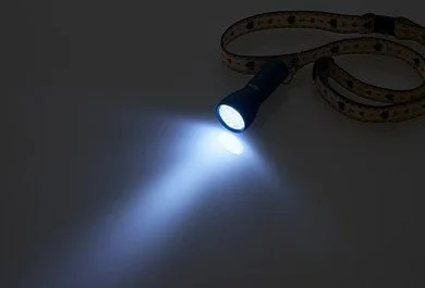 暗い背景に置かれた黒い懐中電灯が、強力な白い光の束を放っている様子。光は暗い表面を照らし、その効果を際立たせている。ストラップも確認できる。
