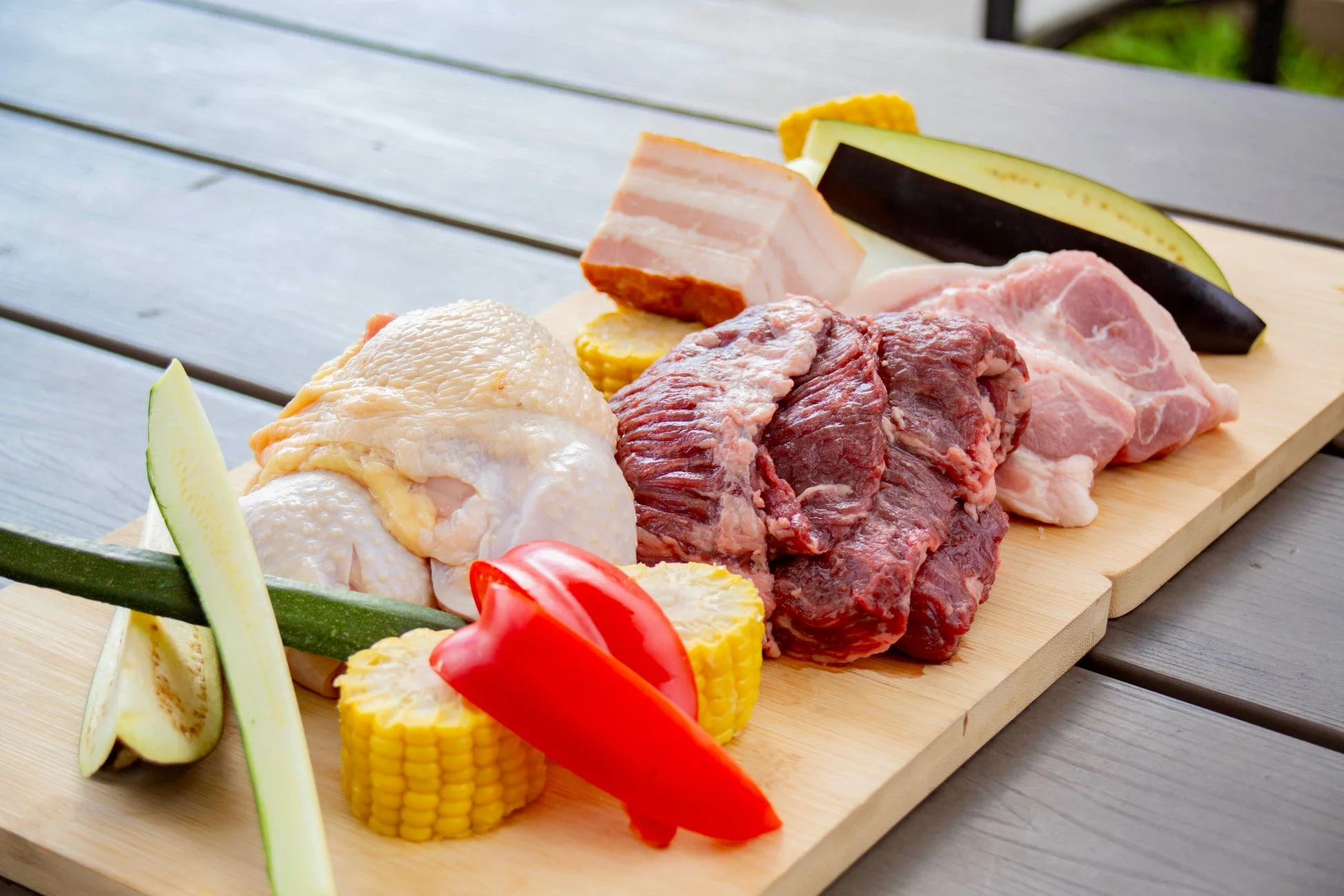 バーベキューの準備として、鶏肉、牛肉、豚肉の様々な部位と、トウモロコシ、パプリカ、ナス、ズッキーニなどの新鮮な野菜が木製のカッティングボードに並べられています