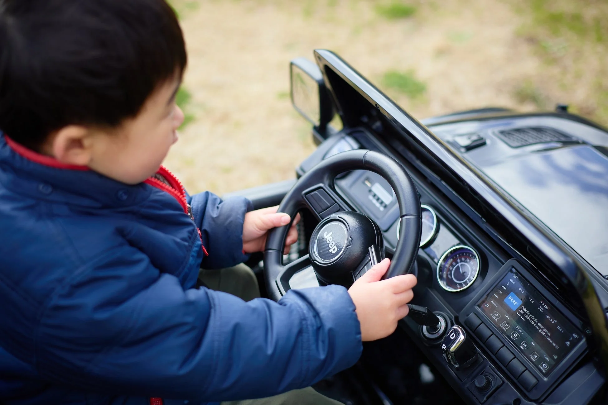 青いジャケットを着た小さな子供が、おもちゃのジープのハンドルを握り、真剣な表情で運転している様子を捉えた画像です。